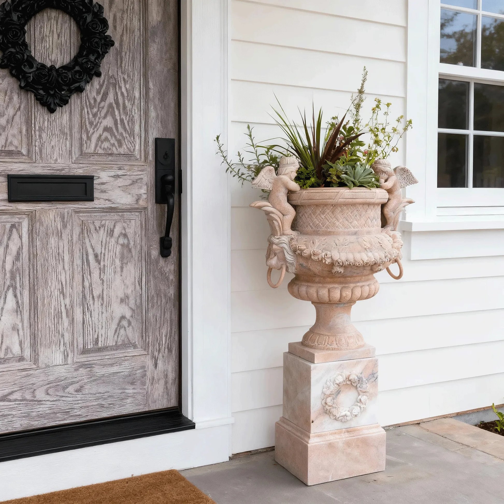 Marble planter sculpture with angel figures, lion head carvings, and floral relief base, displayed at a villa entrance with plants inside.