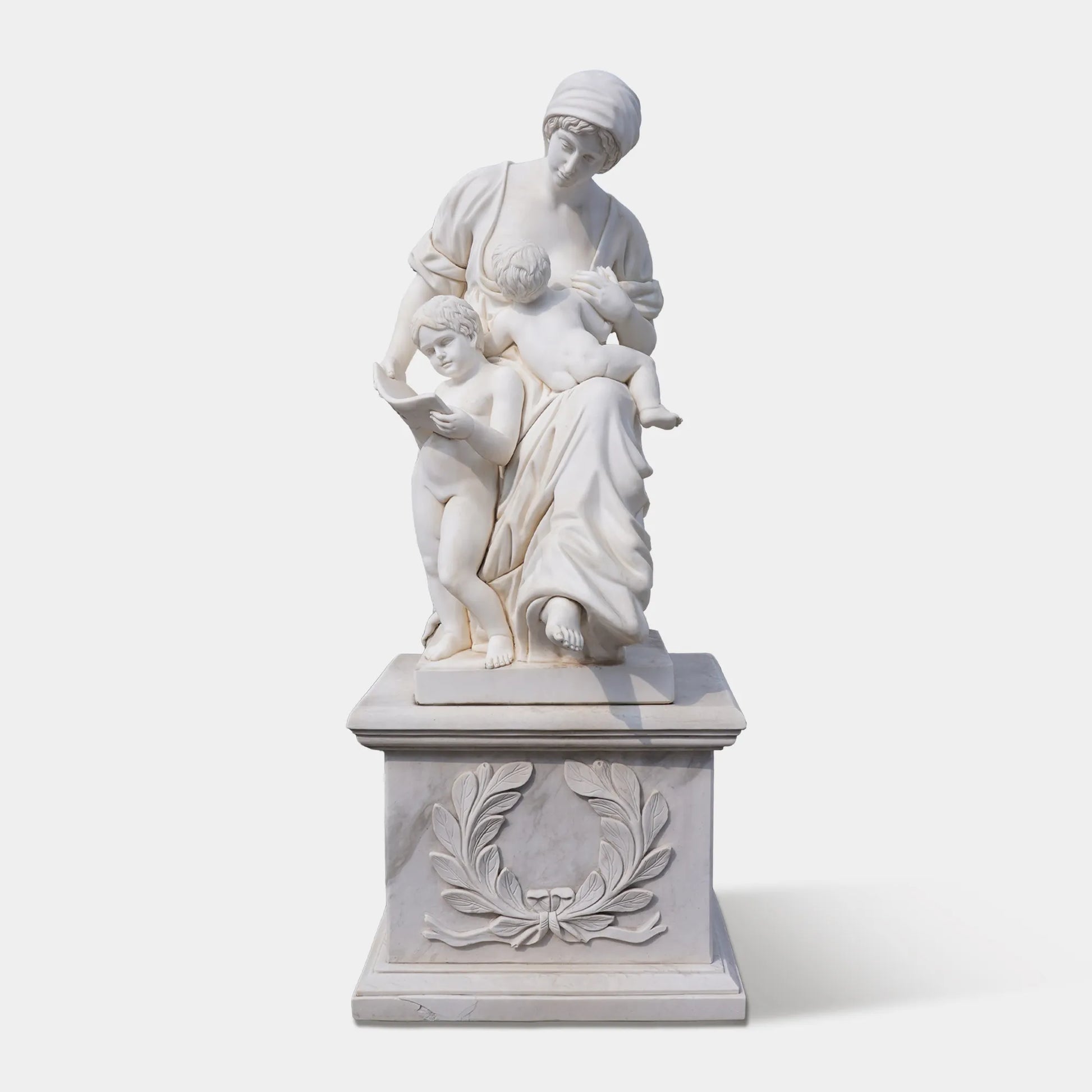 Marble sculpture of a mother with two children reading together on a carved pedestal, displayed outdoors in a European plaza near a historic building.