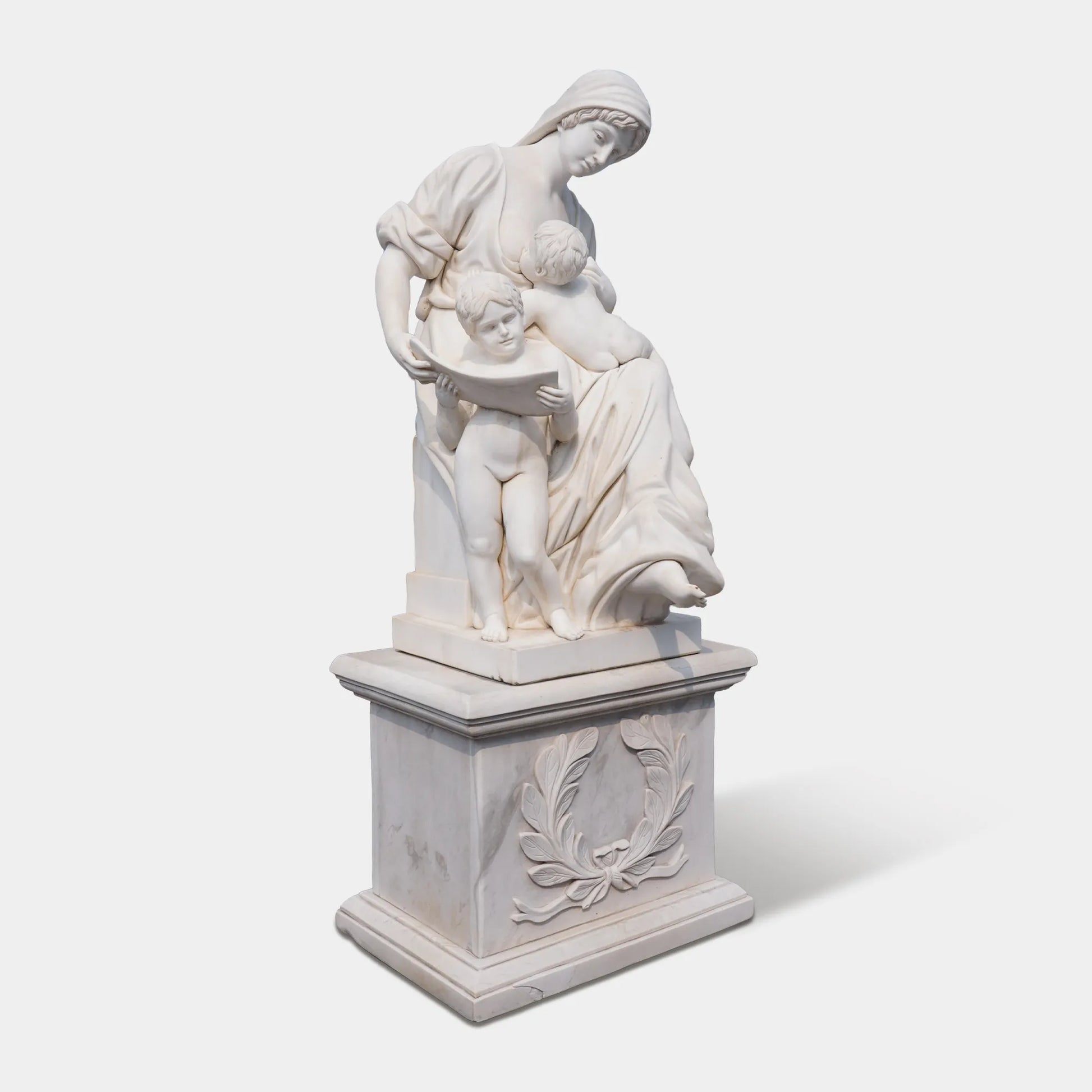 Marble sculpture of a mother with two children reading together on a carved pedestal, displayed outdoors in a European plaza near a historic building.