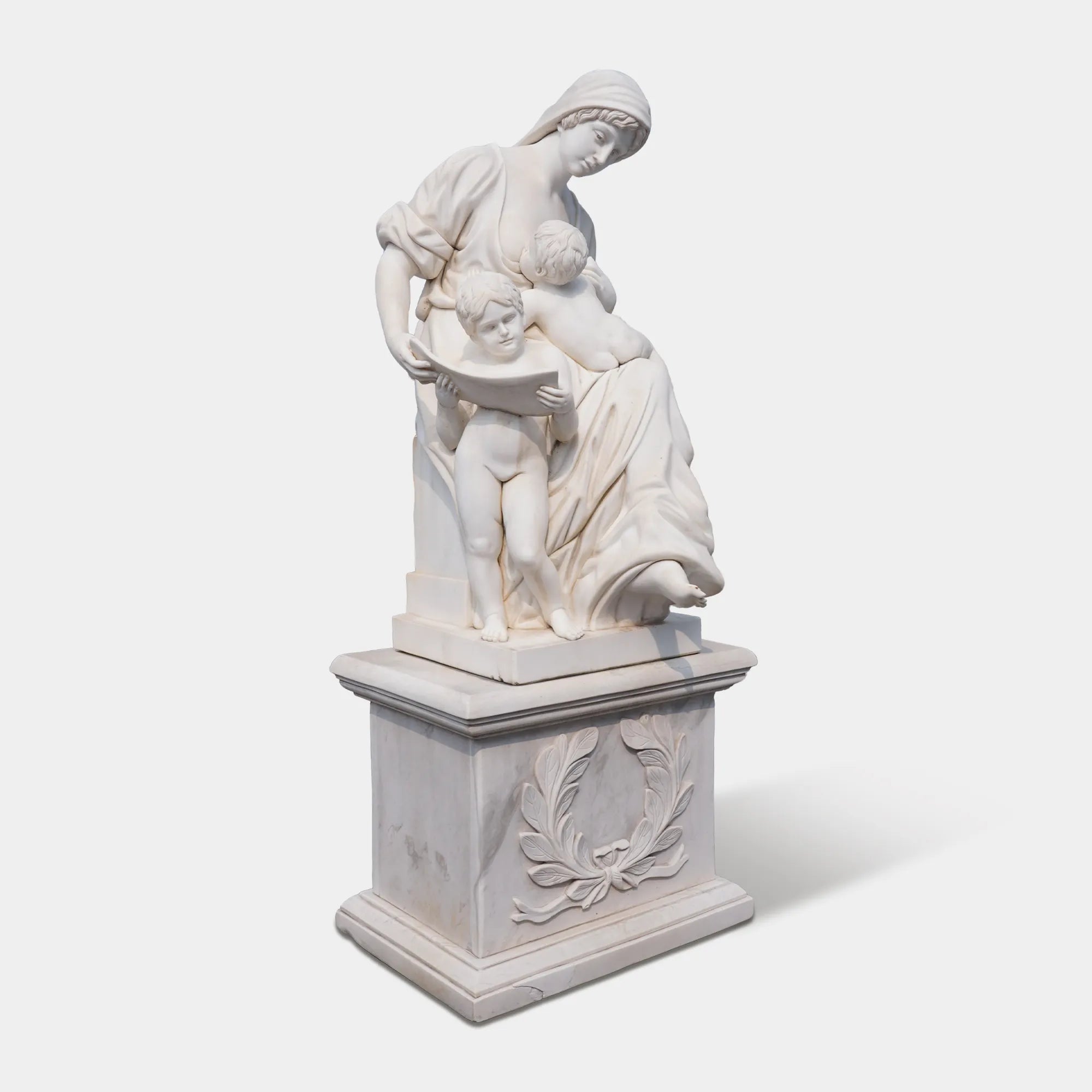Marble sculpture of a mother with two children reading together on a carved pedestal, displayed outdoors in a European plaza near a historic building.