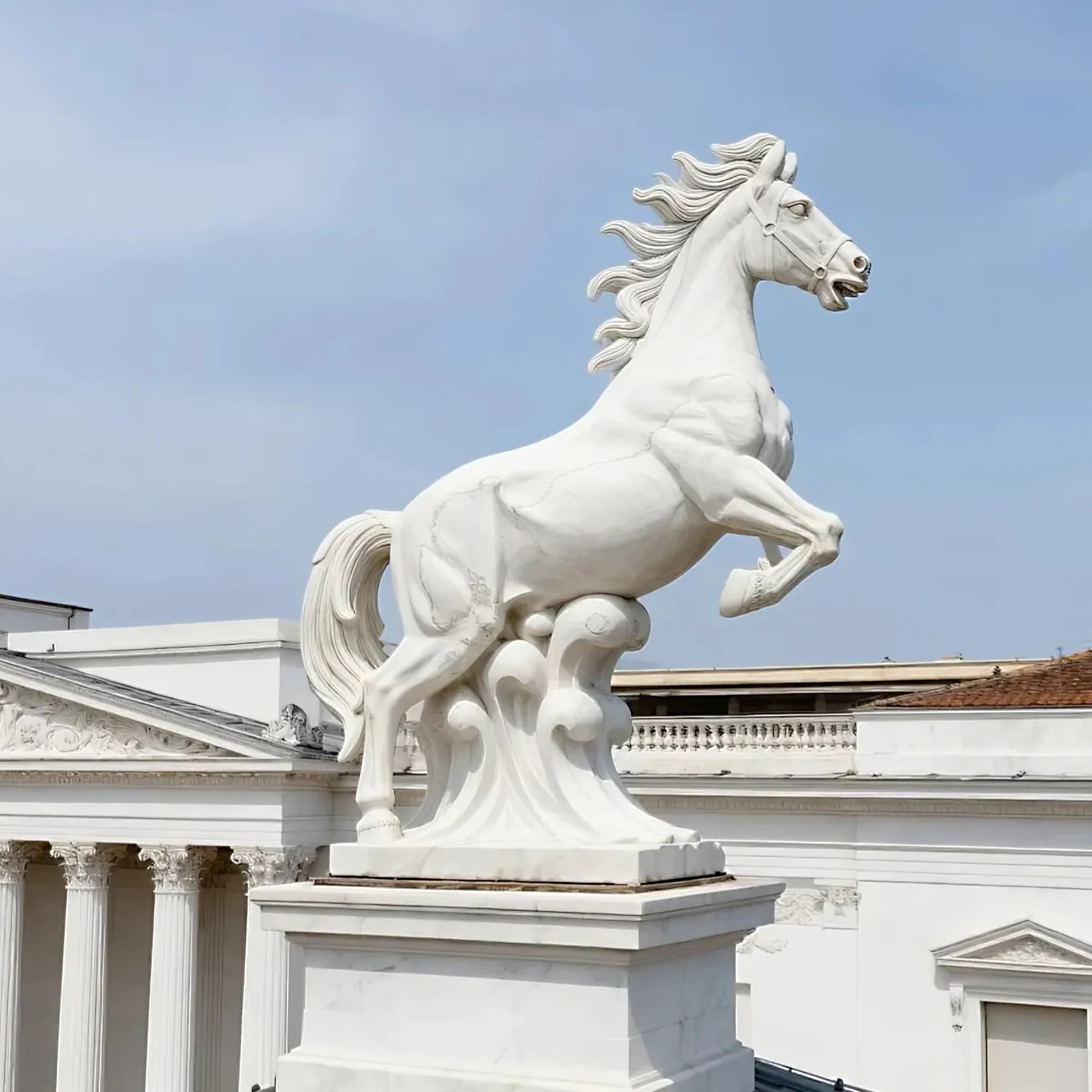 Marble horse sculpture displayed on a pedestal outside a neoclassical building under clear sky.