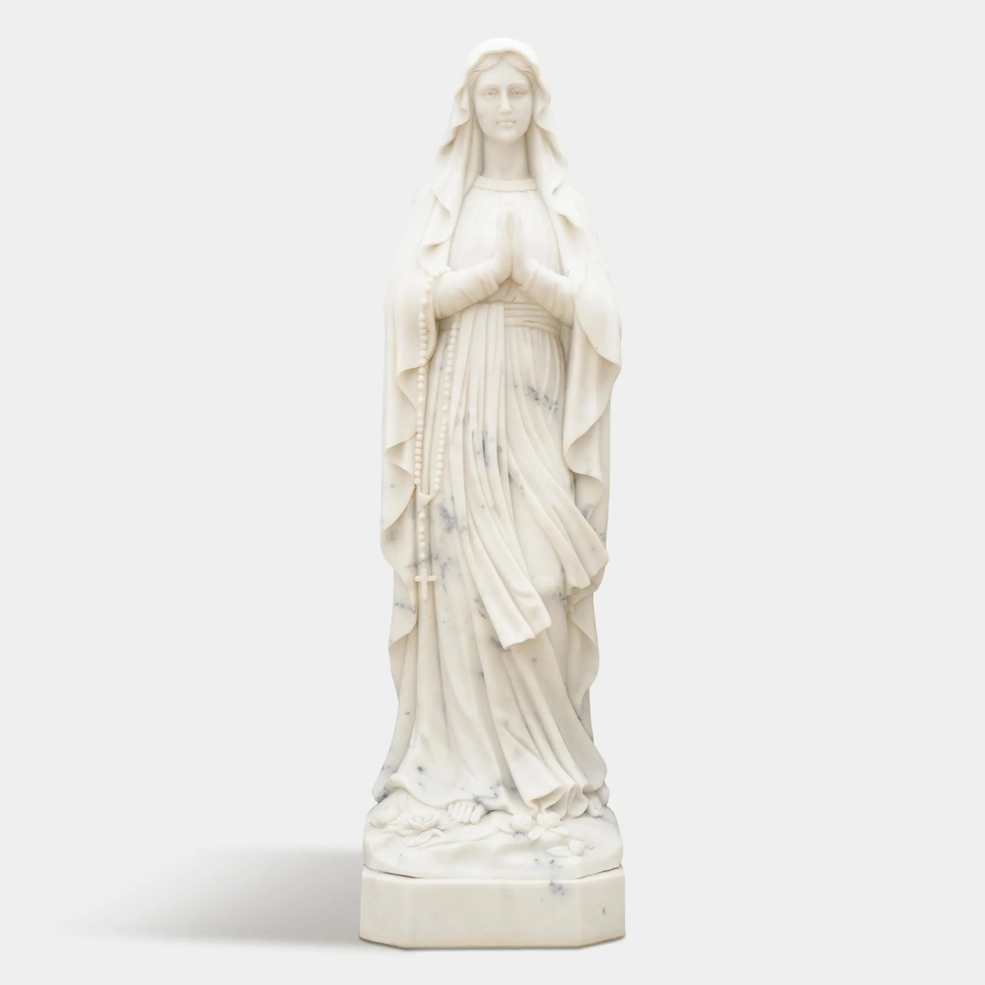 Marble sculpture of Virgin Mary standing in prayer with detailed robe texture displayed in a church or outdoor garden.