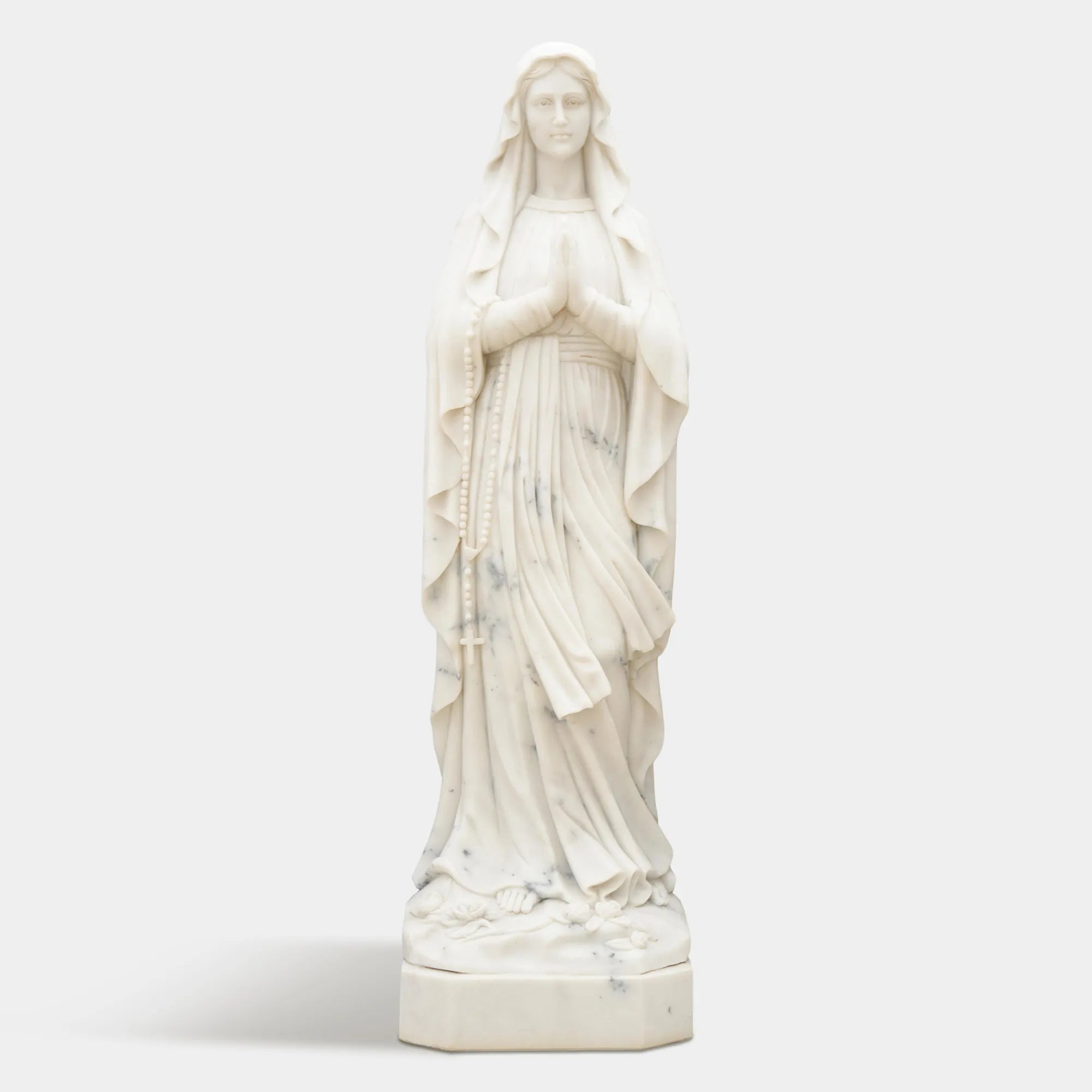 Marble sculpture of Virgin Mary standing in prayer with detailed robe texture displayed in a church or outdoor garden.
