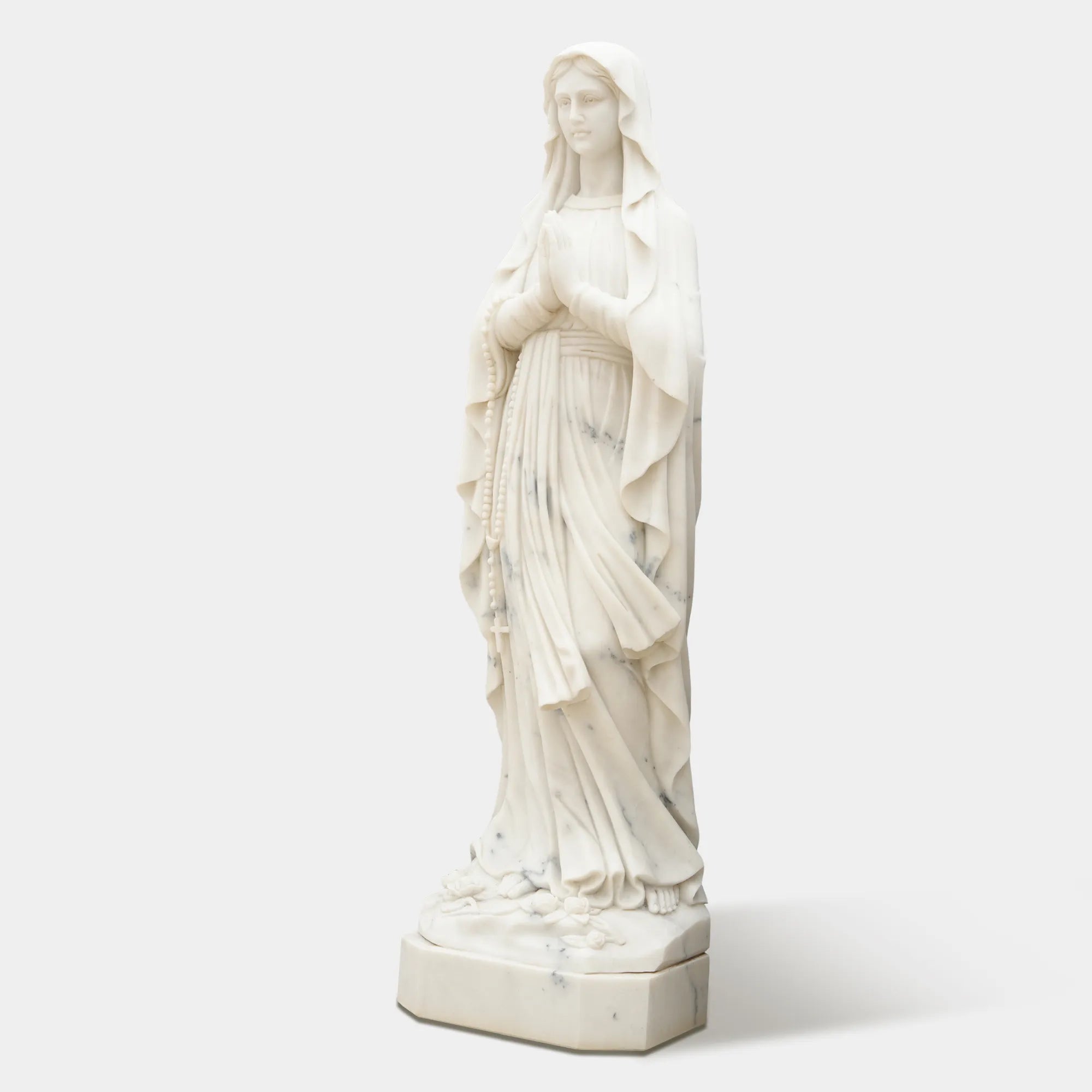 Marble sculpture of Virgin Mary standing in prayer with detailed robe texture displayed in a church or outdoor garden.