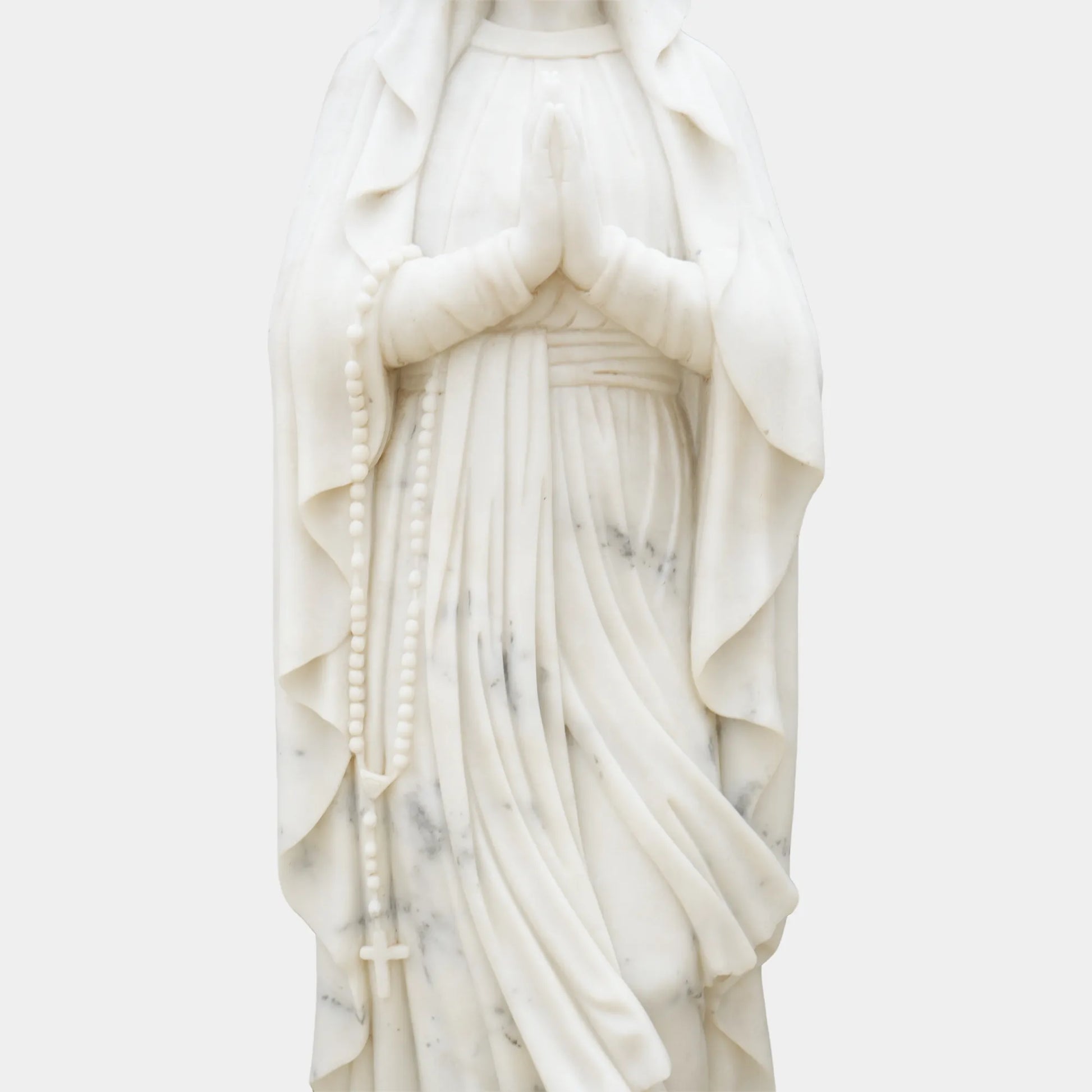 Marble sculpture of Virgin Mary standing in prayer with detailed robe texture displayed in a church or outdoor garden.
