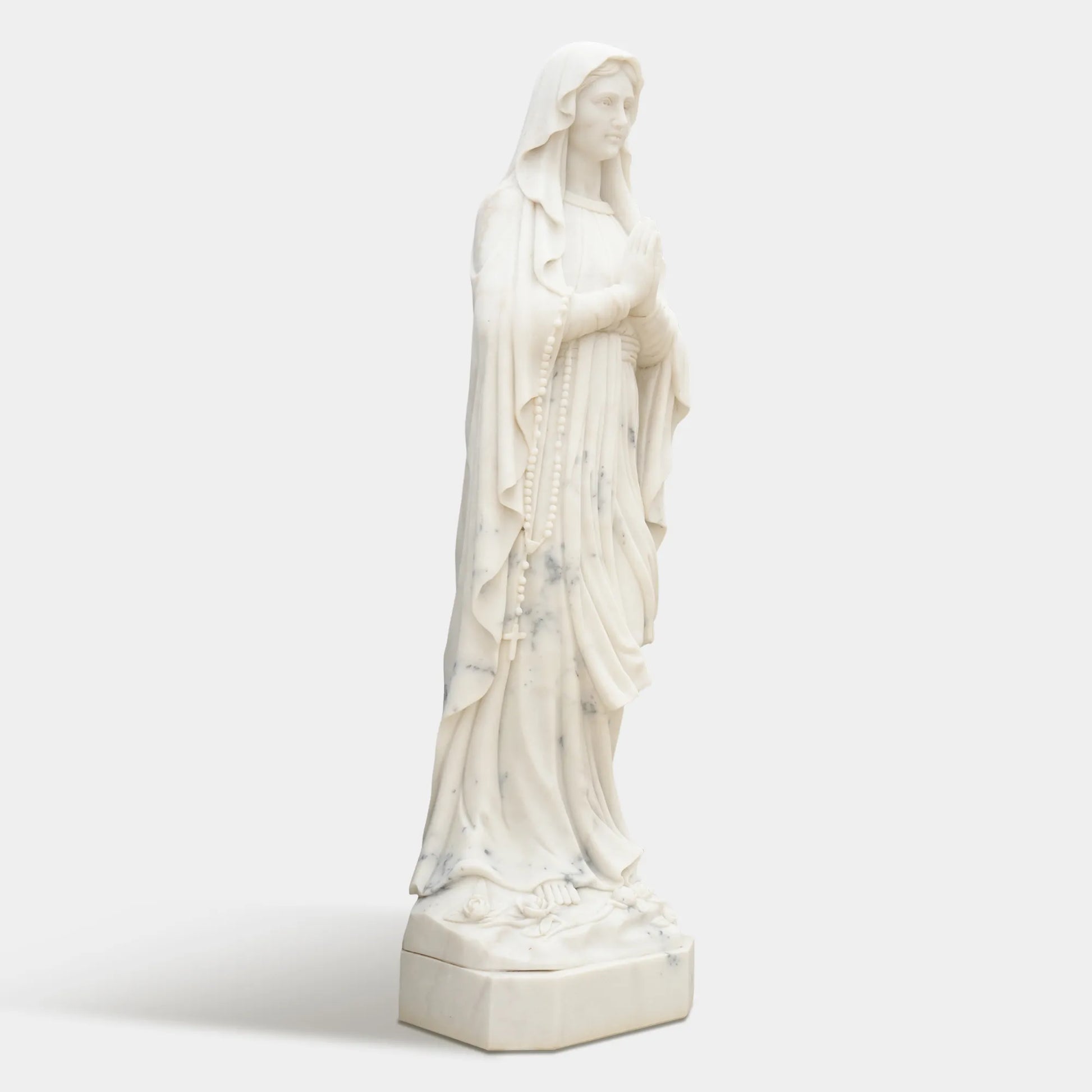 Marble sculpture of Virgin Mary standing in prayer with detailed robe texture displayed in a church or outdoor garden.