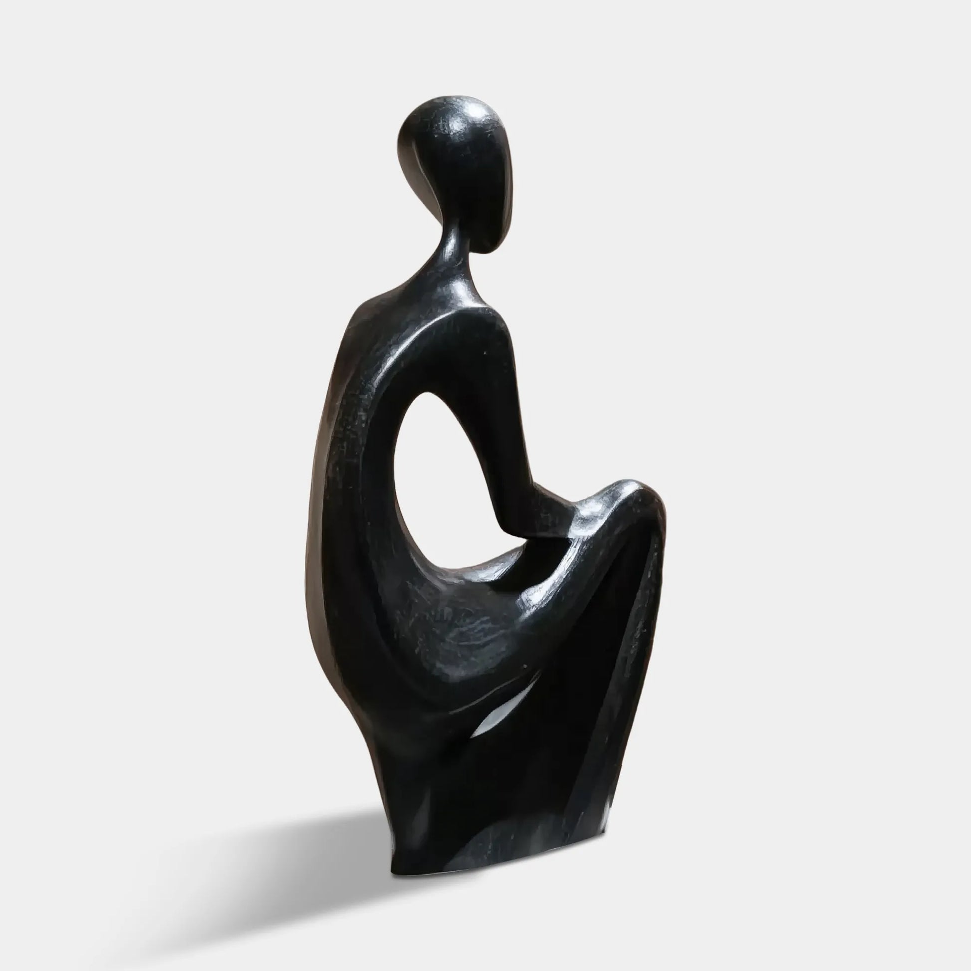 Black bronze abstract seated figurative sculpture on a pedestal in a modern glass-walled gallery space. 
