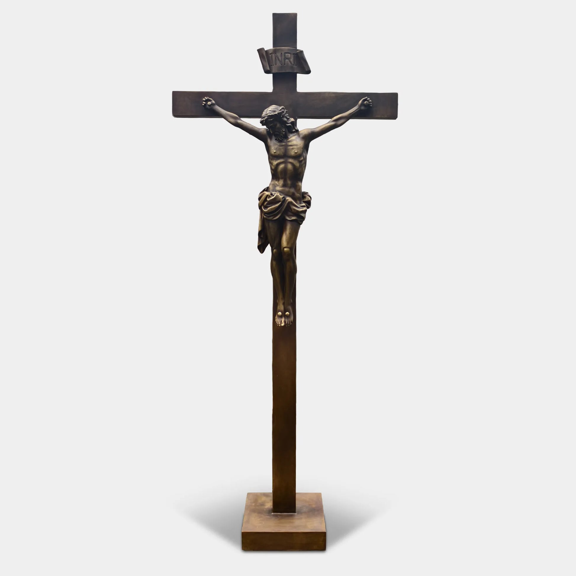 Bronze crucifix religious statue with Jesus figure mounted on a cross, displayed outdoors on a lawn between two tall evergreen trees.