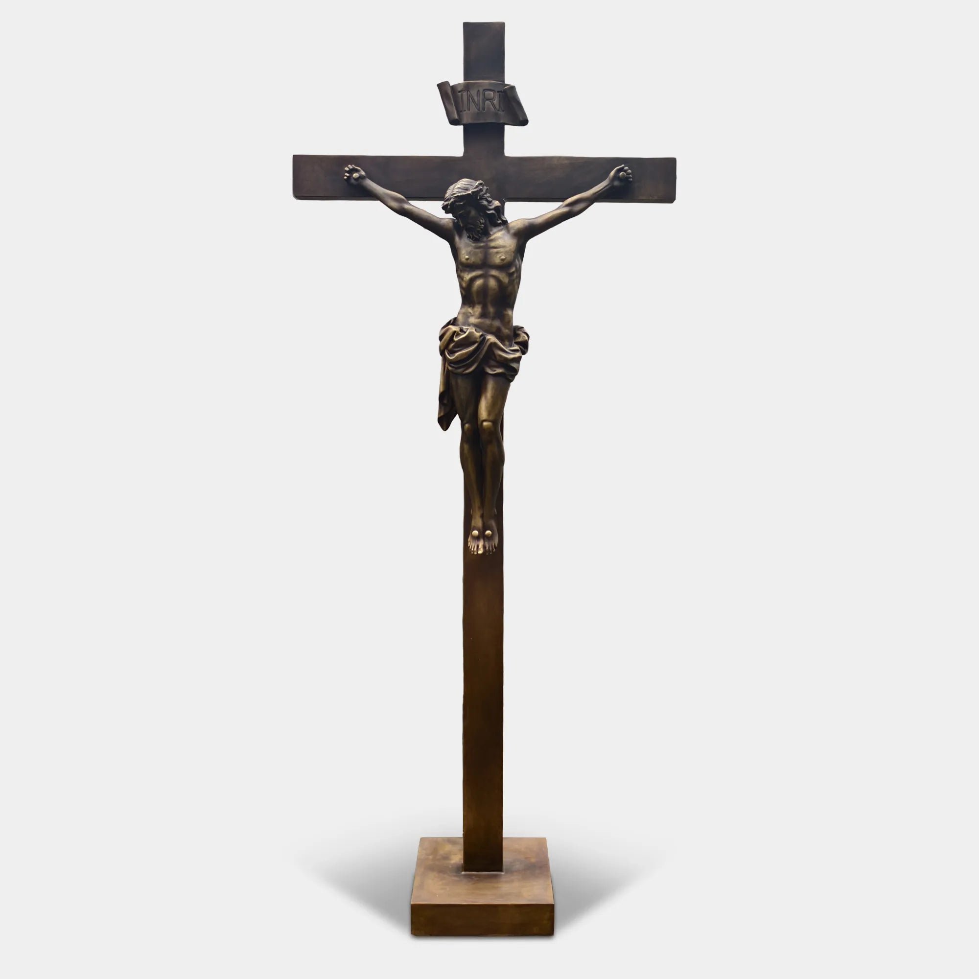 Bronze crucifix religious statue with Jesus figure mounted on a cross, displayed outdoors on a lawn between two tall evergreen trees.