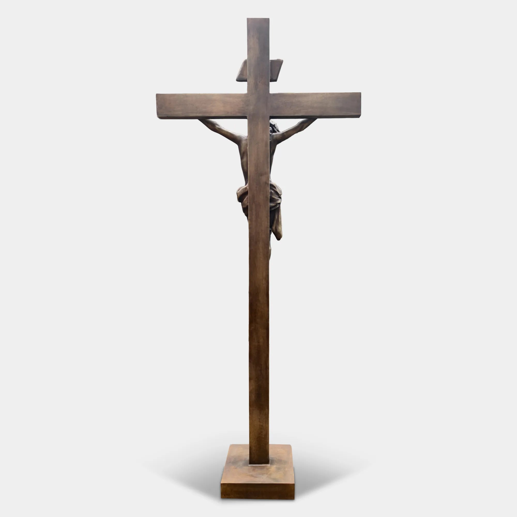 Bronze crucifix religious statue with Jesus figure mounted on a cross, displayed outdoors on a lawn between two tall evergreen trees.