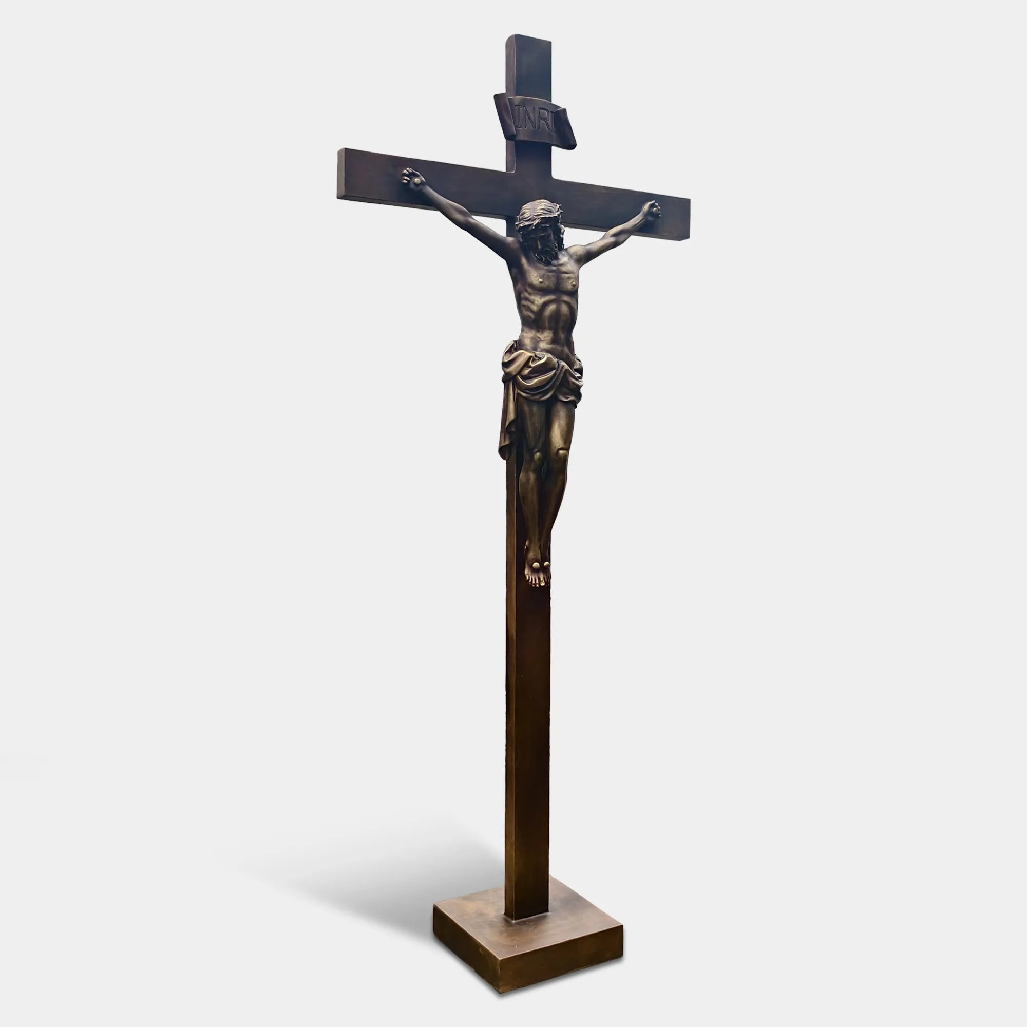 Bronze crucifix religious statue with Jesus figure mounted on a cross, displayed outdoors on a lawn between two tall evergreen trees.