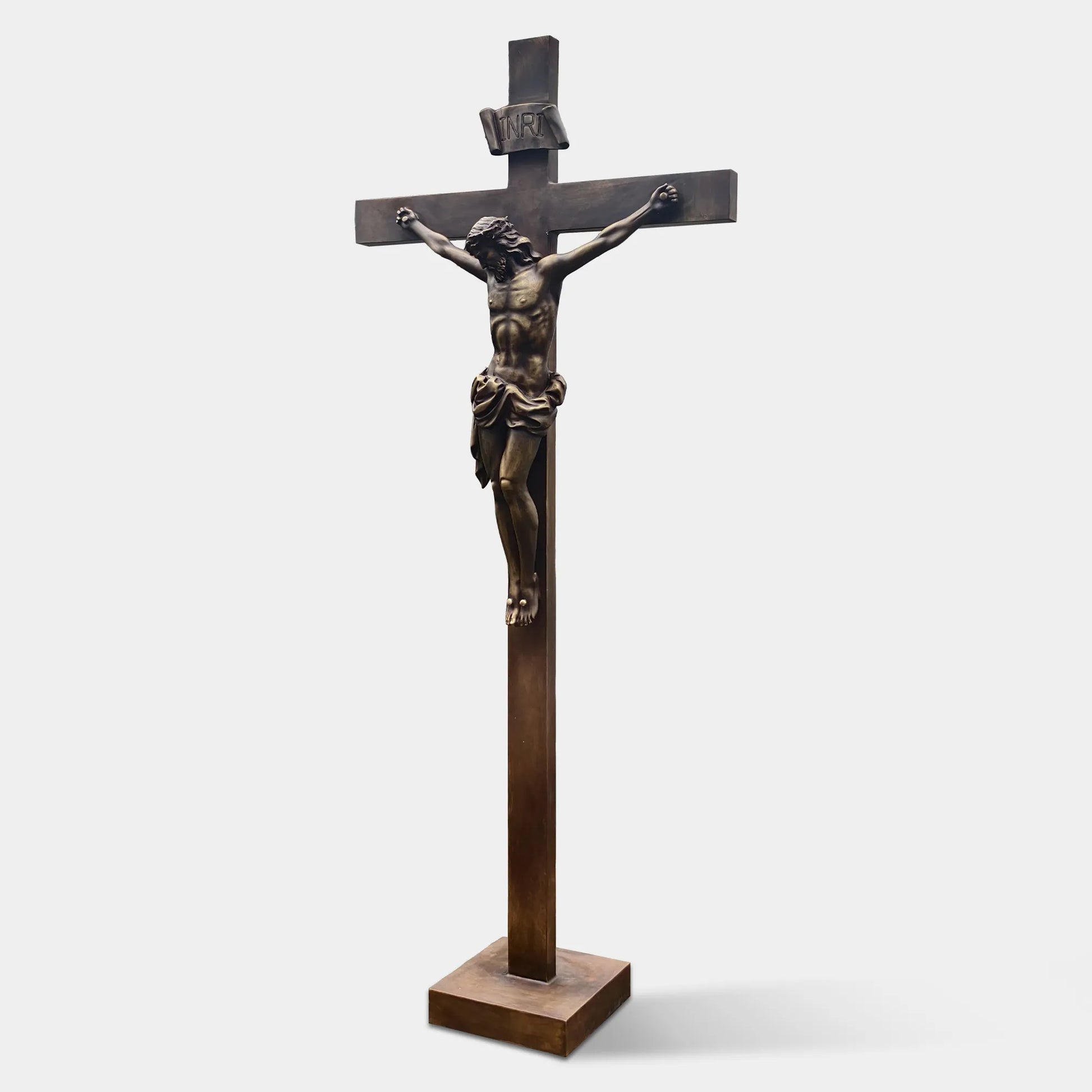 Bronze crucifix religious statue with Jesus figure mounted on a cross, displayed outdoors on a lawn between two tall evergreen trees.