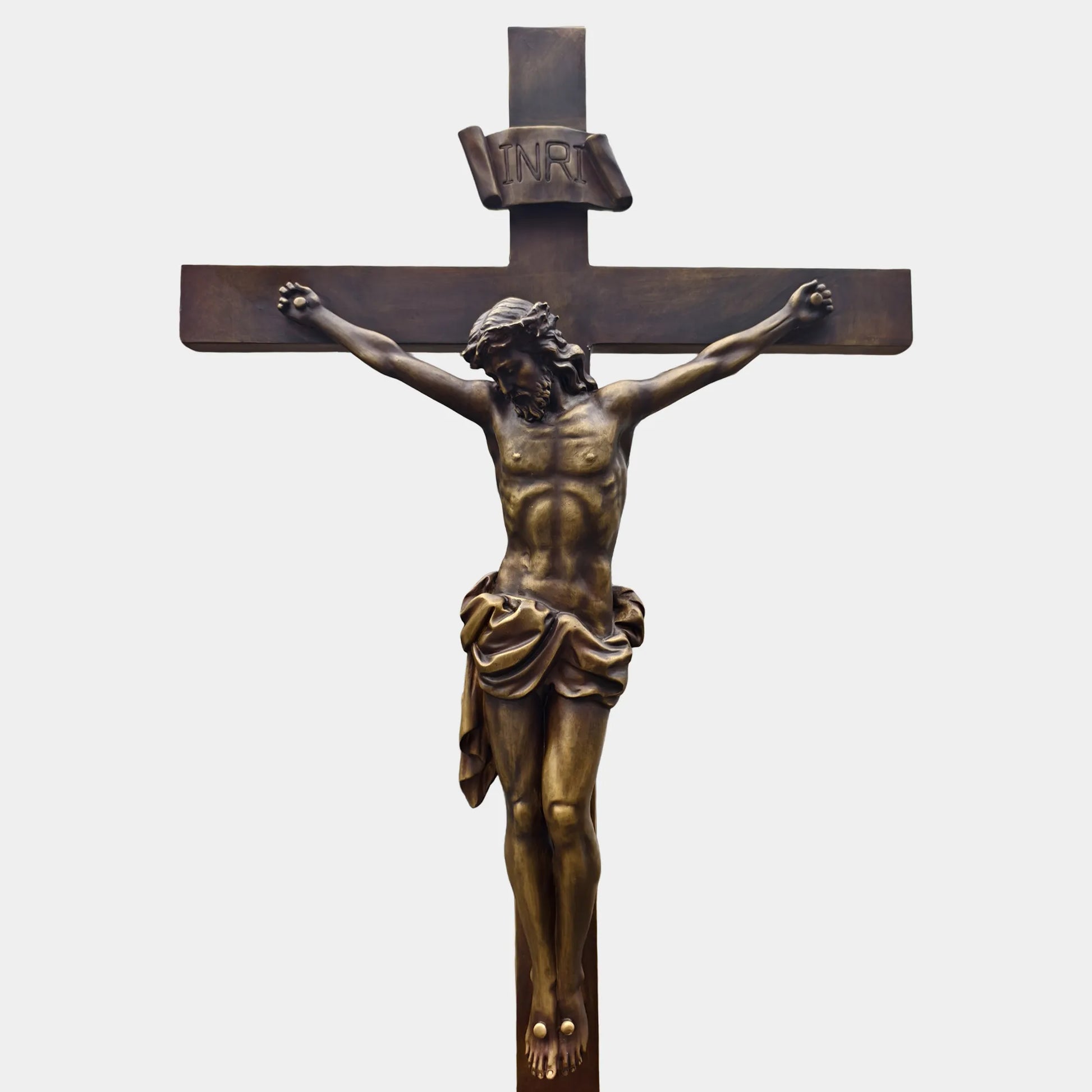 Bronze crucifix religious statue with Jesus figure mounted on a cross, displayed outdoors on a lawn between two tall evergreen trees.