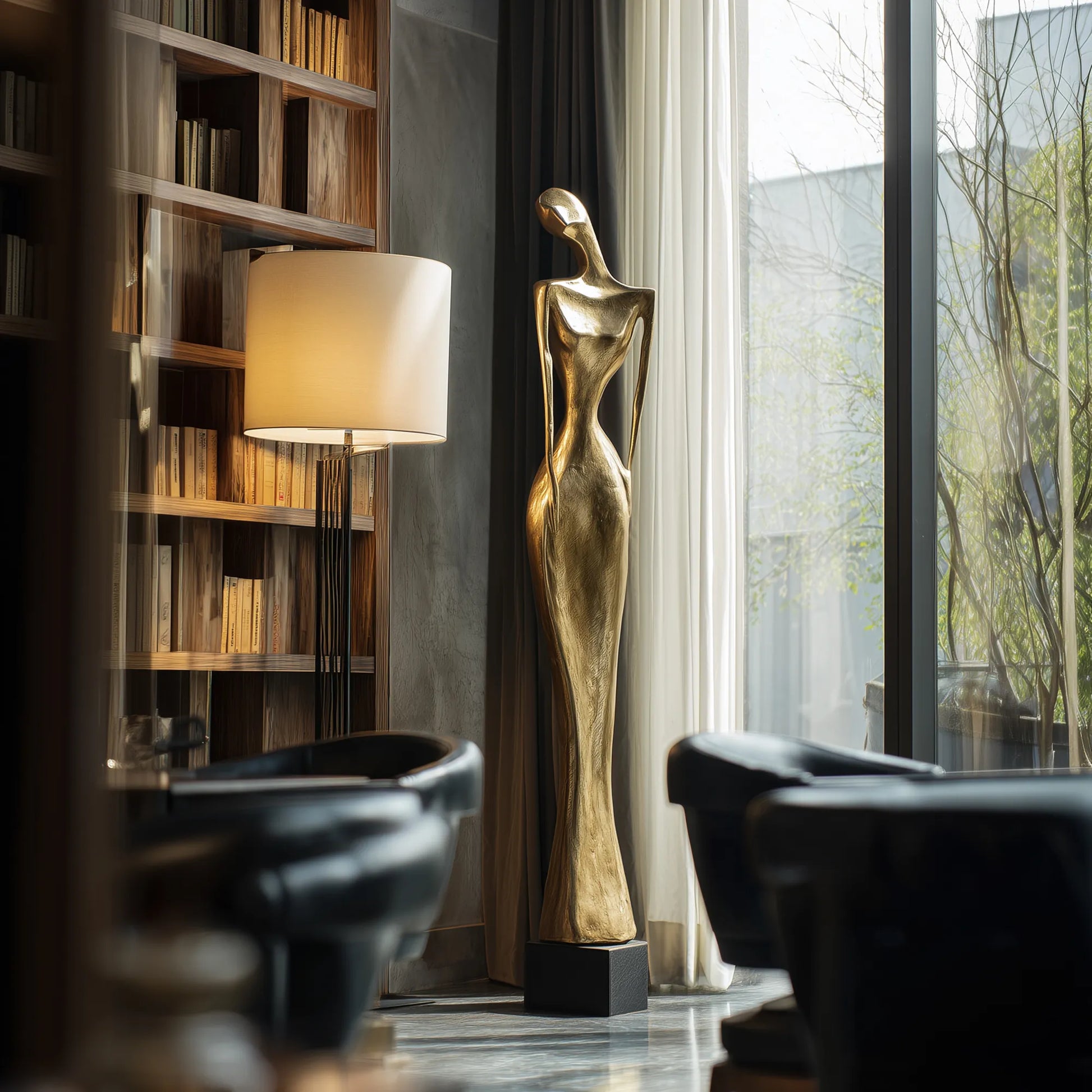 Bronze abstract figurative sculpture on black base beside bookshelf and window. 