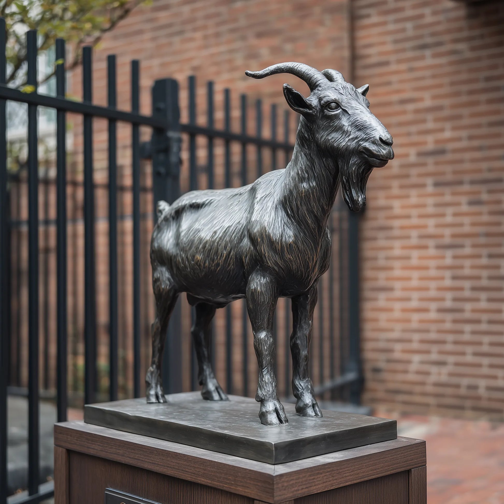 A realistic bronze goat sculpture stands outdoors, with a black iron fence and red brick wall in the background.