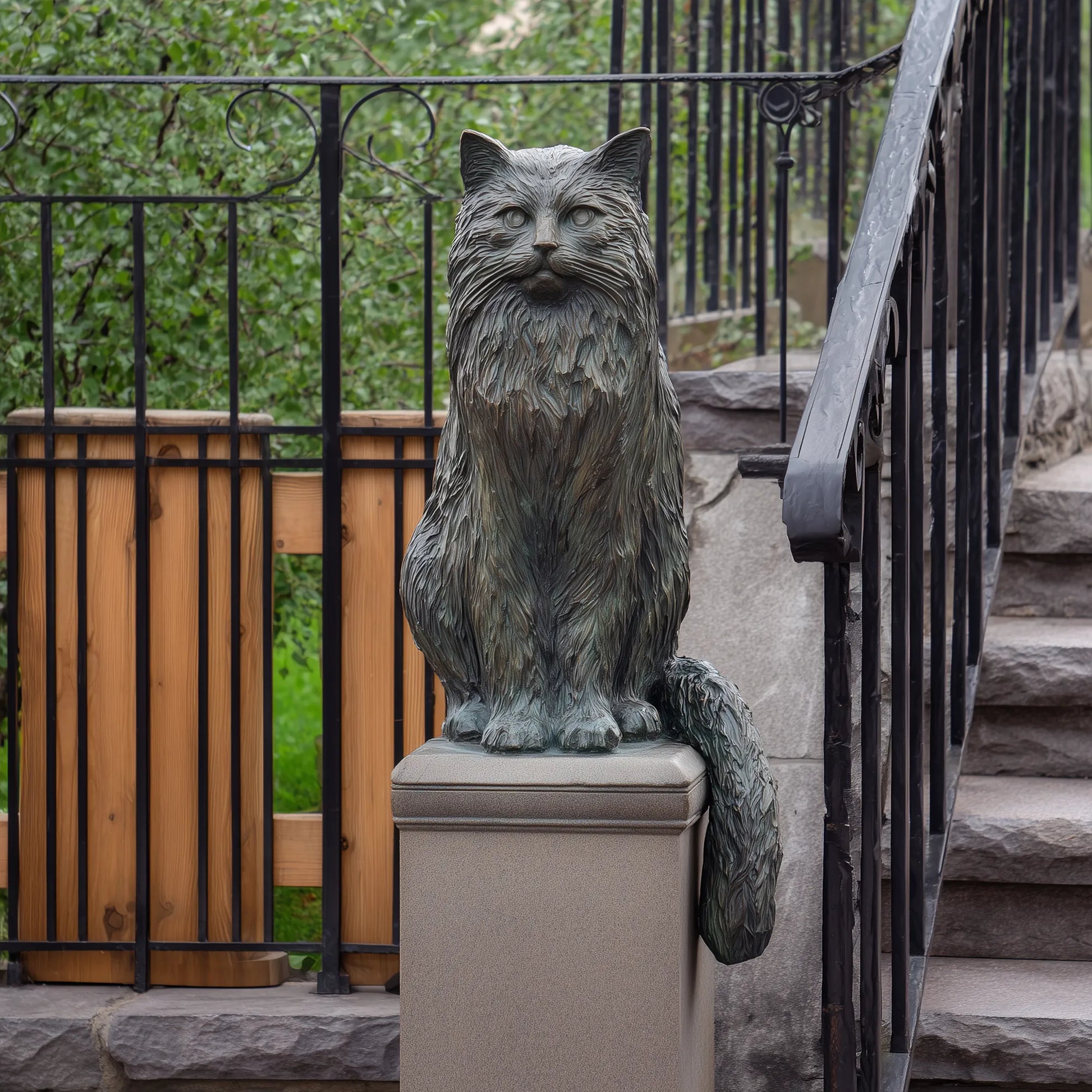 Bronze long haired cat statue sits on porch pillar beside stone steps and black metal fence.
