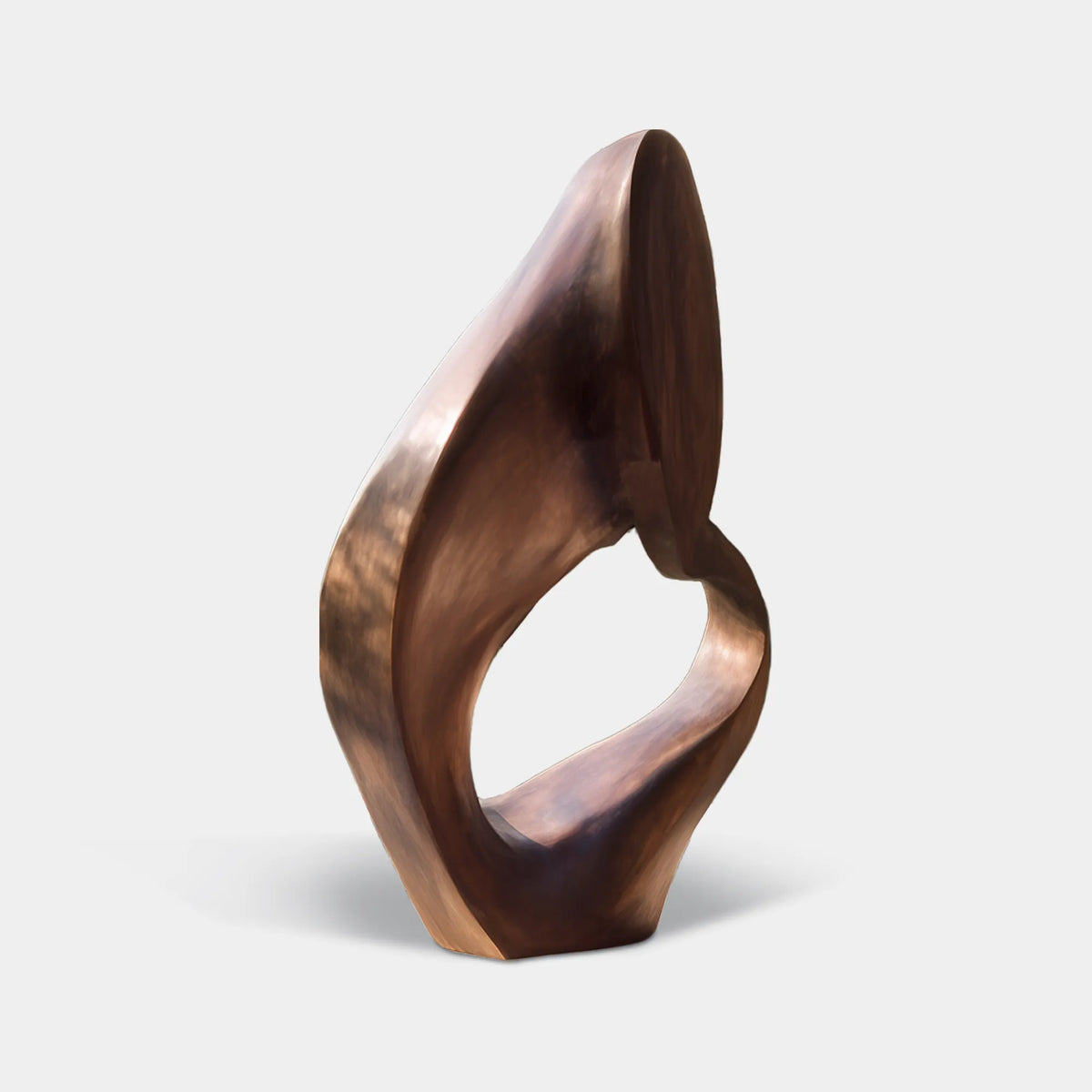 Warm bronze sculpture with a twisted organic silhouette and large open center on a black pedestal in a modern courtyard.