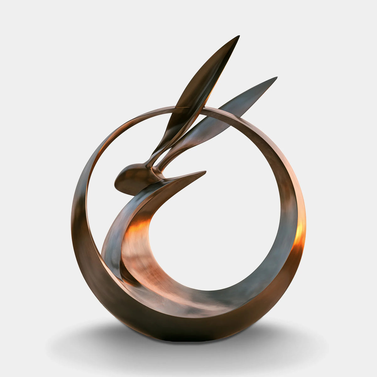 Abstract bronze rabbit sculpture with circular flowing form in an outdoor landscape.