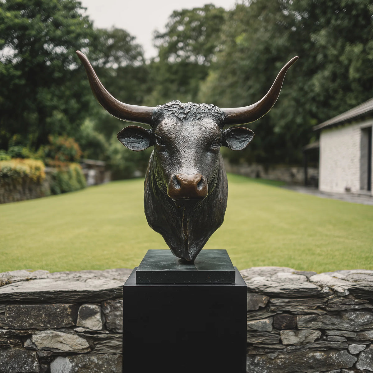 Dark bronze longhorn bull head statue with wide curved horns on a black pedestal in a country estate garden.
