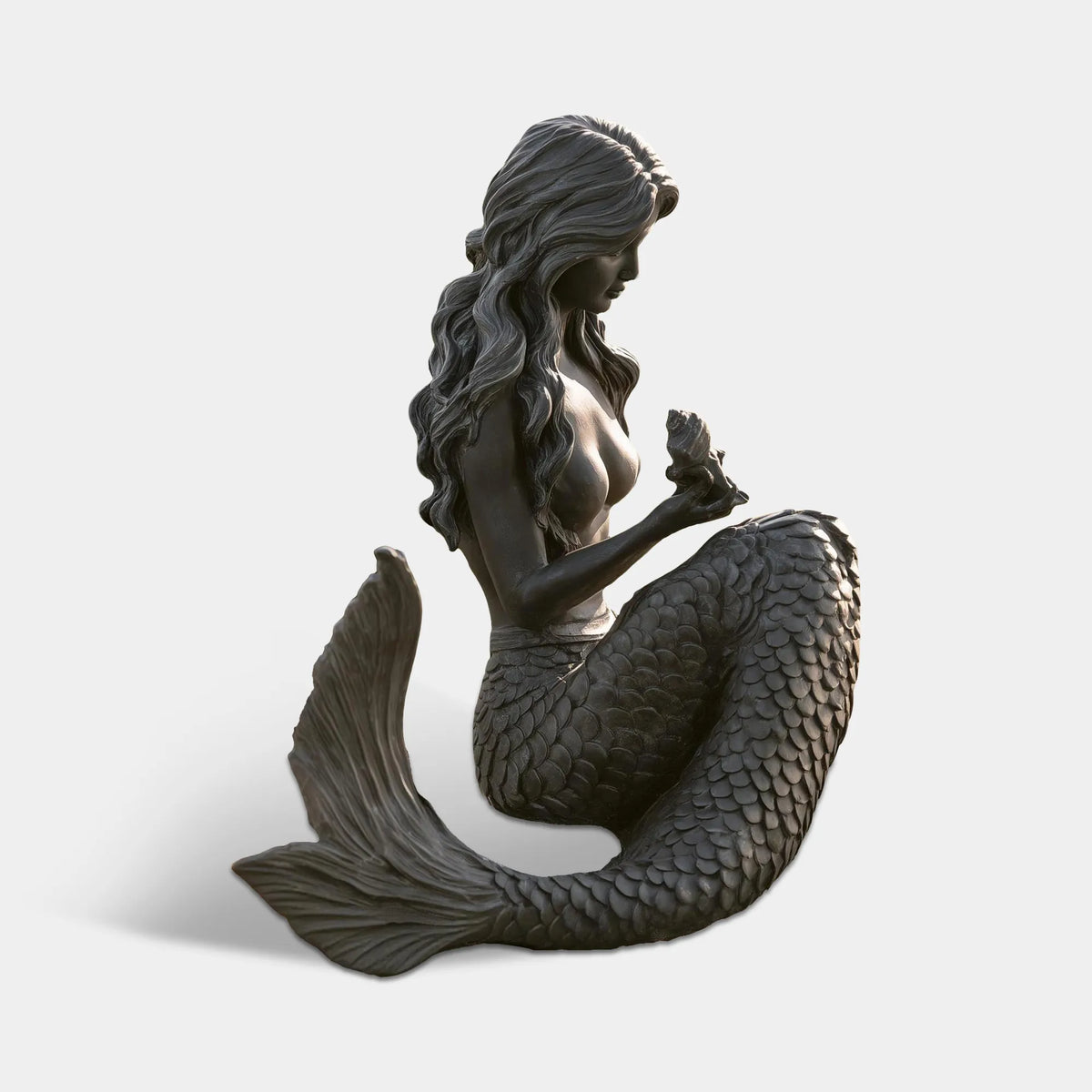 A bronze sitting mermaid statue rests on stacked stones beside a white picket fence.