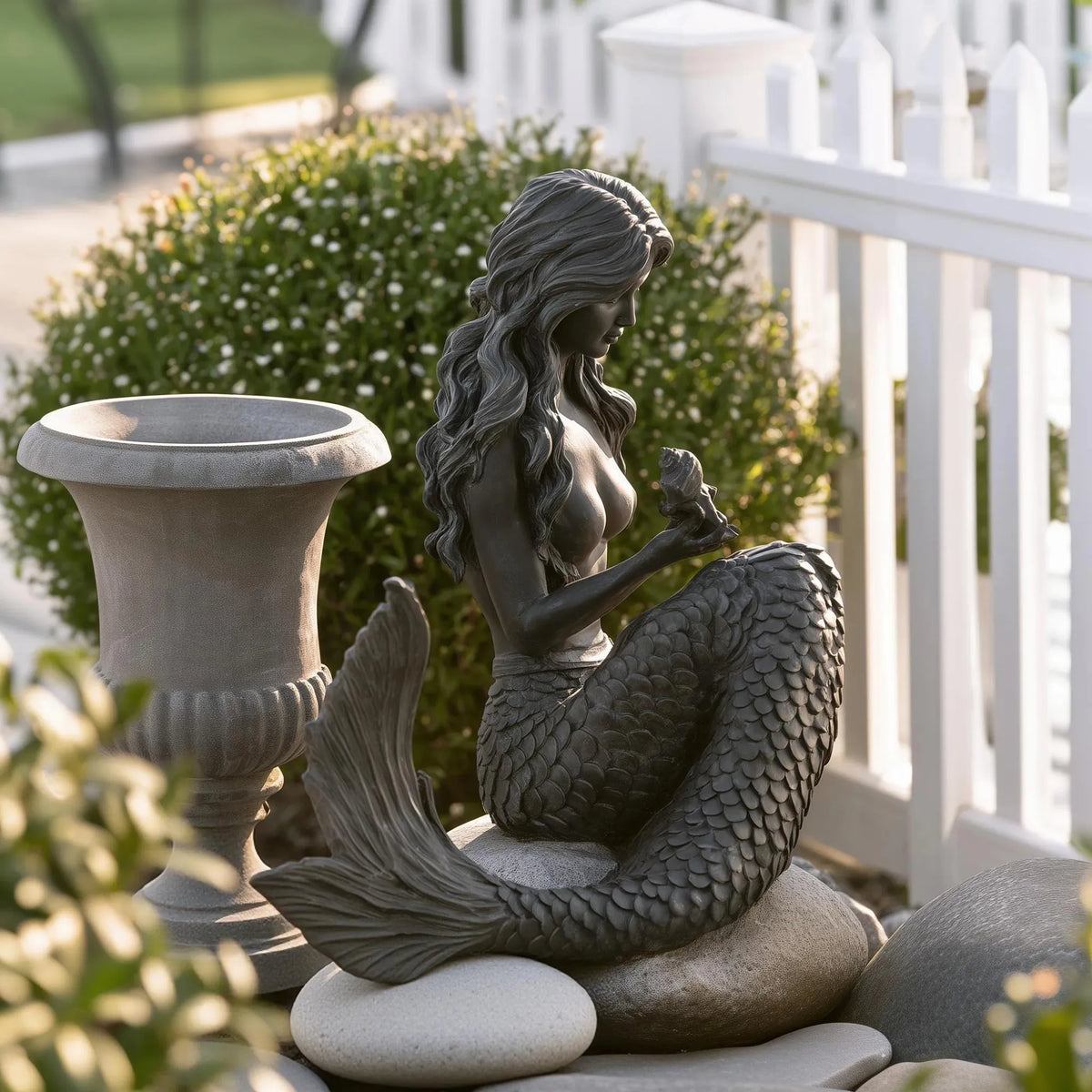 A bronze sitting mermaid statue rests on stacked stones beside a white picket fence.