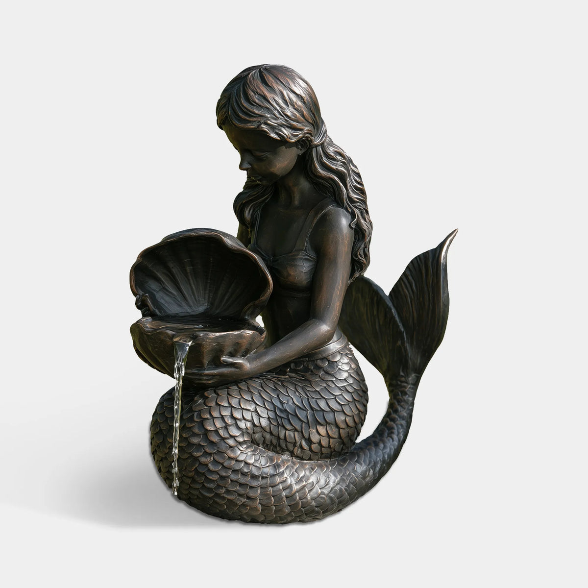A bronze mermaid statue holds a seashell with water flowing out in a green yard.