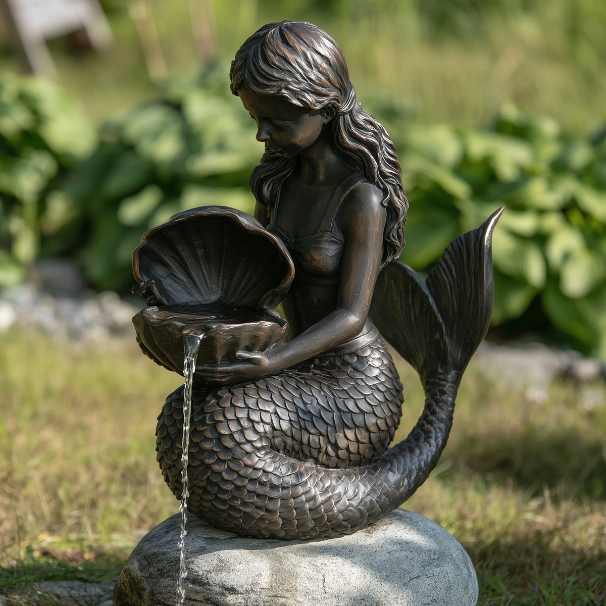 A bronze mermaid statue holds a seashell with water flowing out in a green yard.