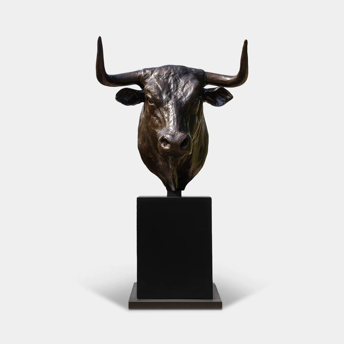 Dark bronze longhorn bull bust sculpture with wide upward-curving horns, realistic facial features and polished highlights on a tall black pedestal outside an elegant building entrance