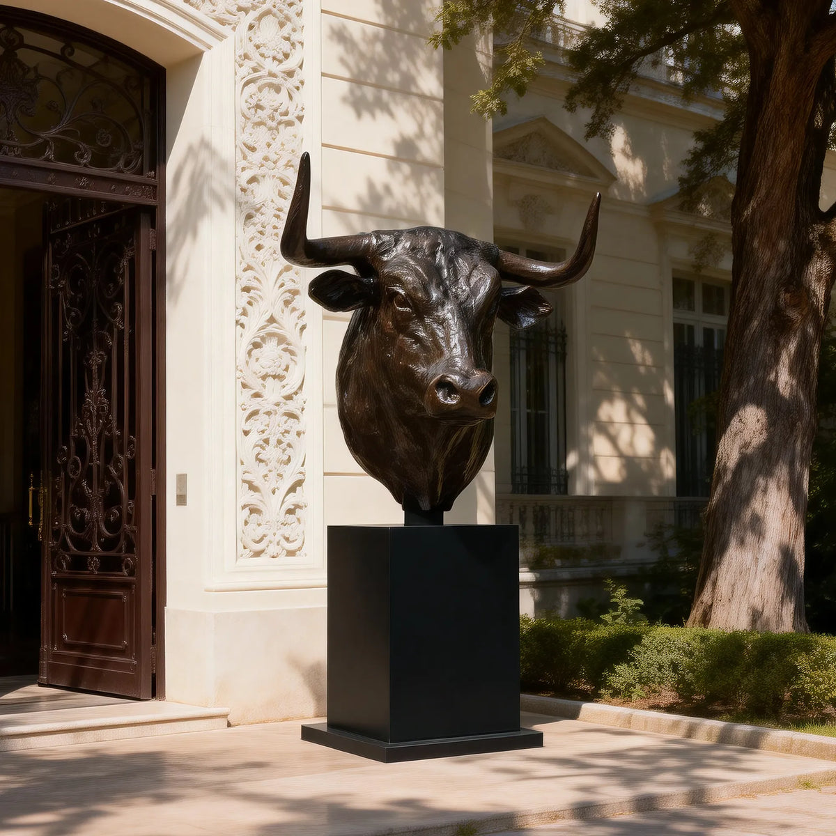 Dark bronze longhorn bull bust sculpture with wide upward-curving horns, realistic facial features and polished highlights on a tall black pedestal outside an elegant building entrance