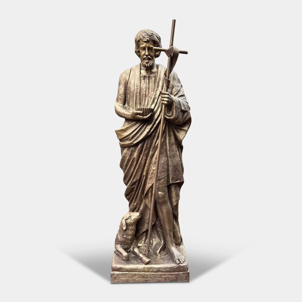 A bronze Saint John the Baptist statue stands on a pedestal in a city park.