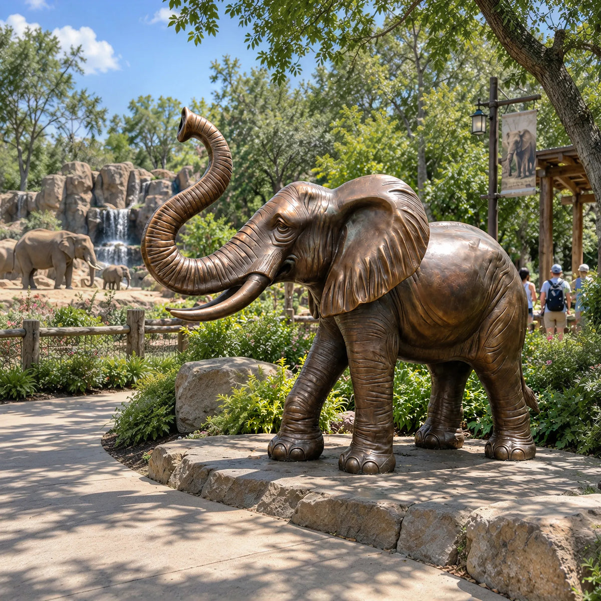 A bronze elephant statue with raised trunk stands along a shaded park walking path.