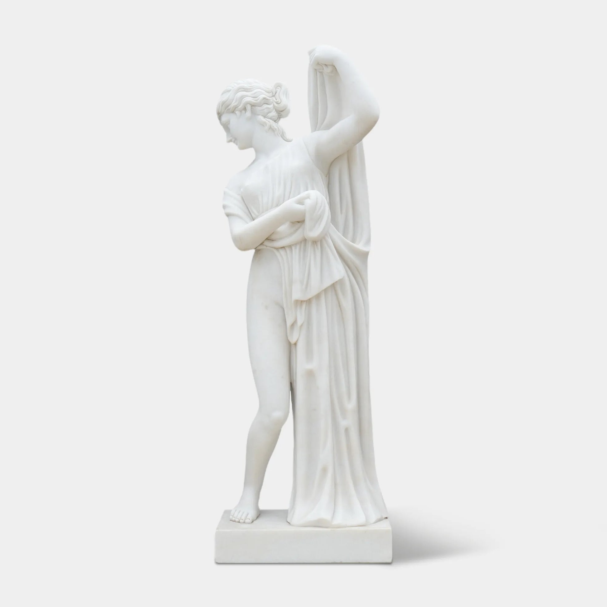 Marble statue of a woman wearing a long dress, gently leaning forward with subtle movement, standing among greenery and flowers.