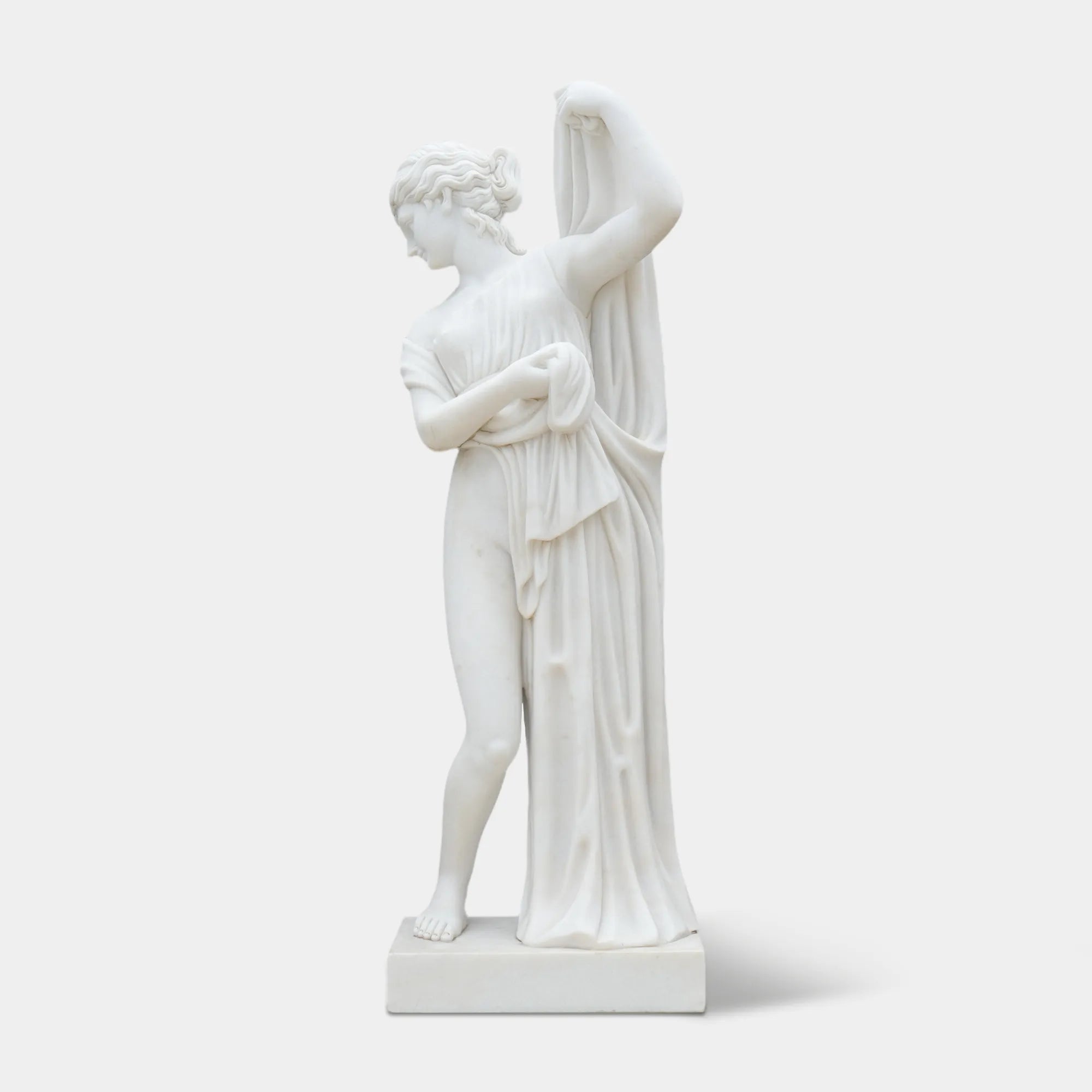 Marble statue of a woman wearing a long dress, gently leaning forward with subtle movement, standing among greenery and flowers.