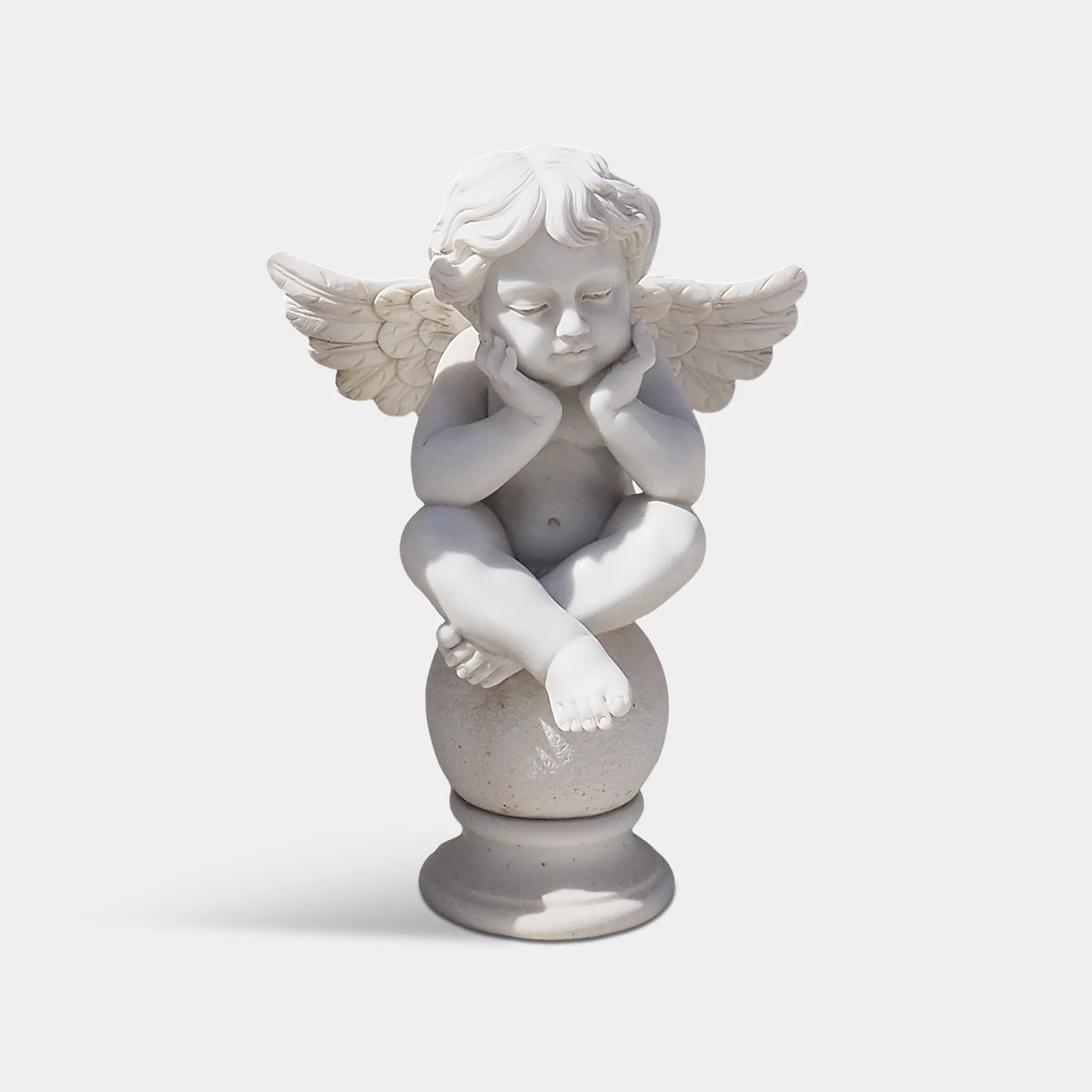 Small angel statue made of natural white marble, sitting on a round pedestal, resting its face on its hands, surrounded by garden greenery