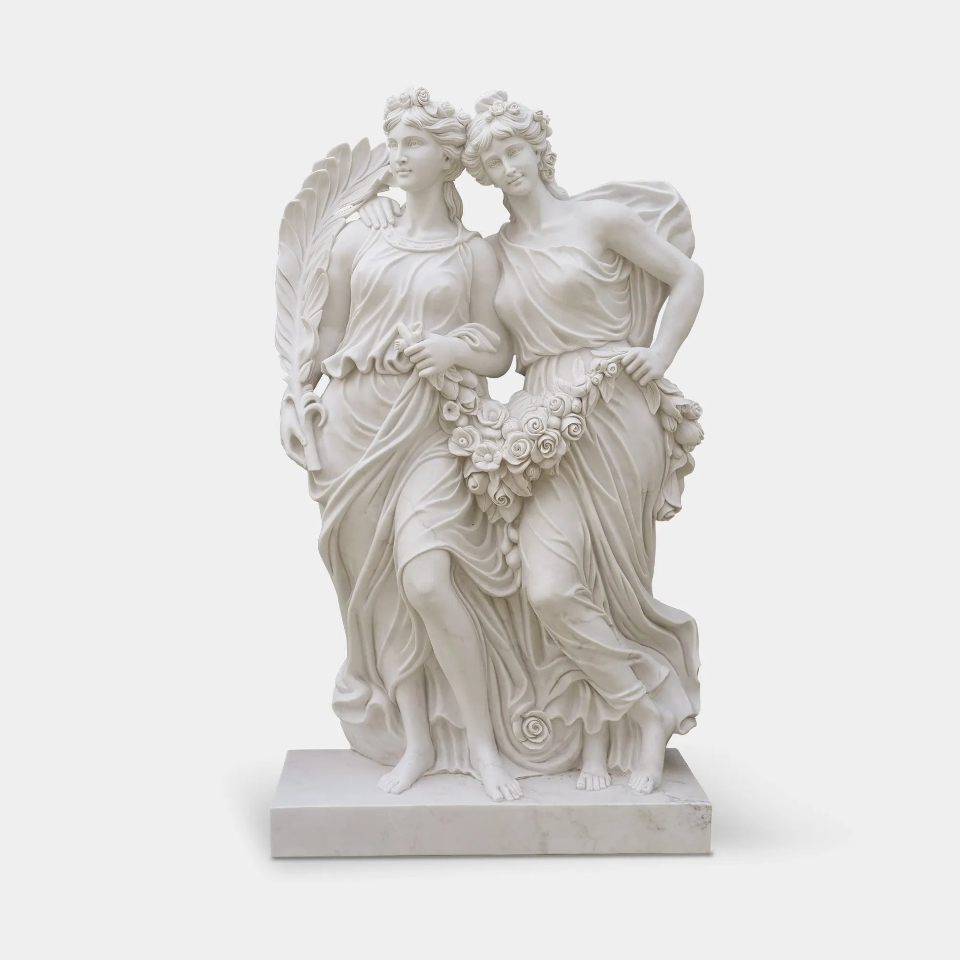 White marble statue of two female figures holding flowers in front of blooming rose garden with classical courtyard behind. 