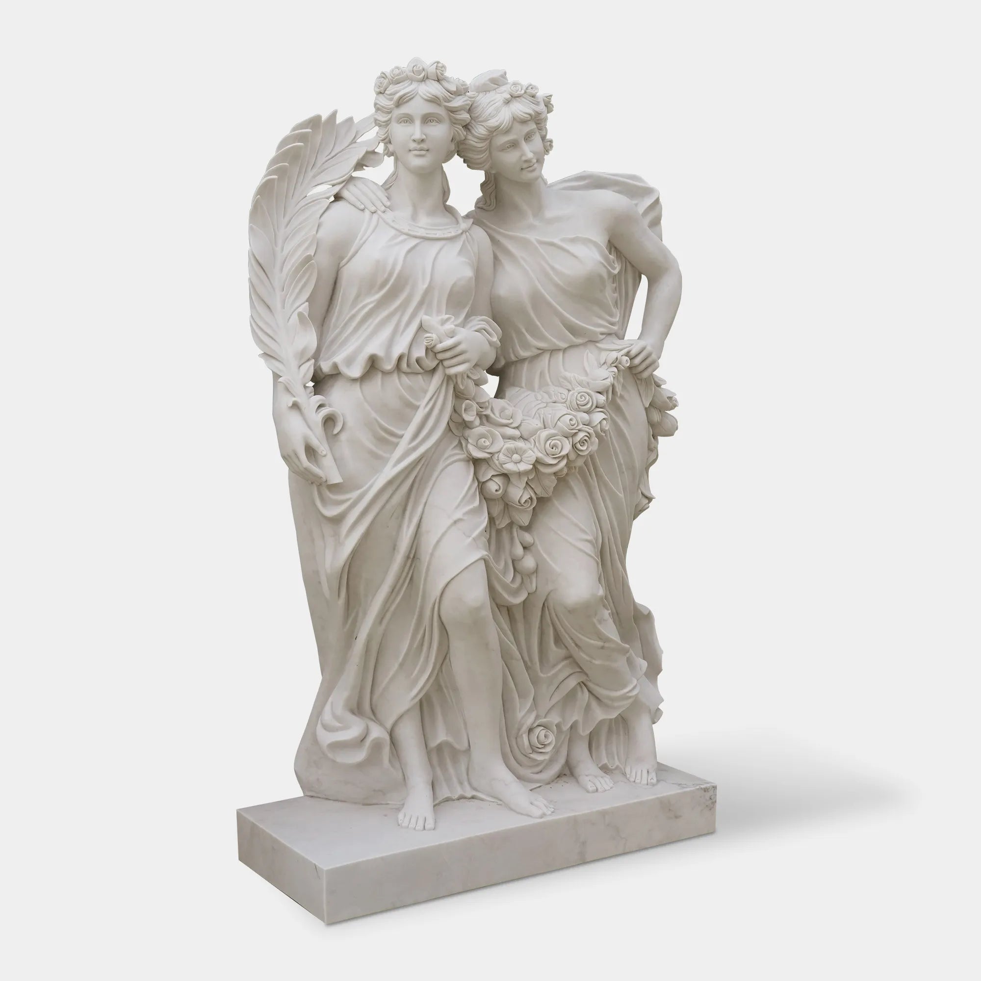 White marble statue of two female figures holding flowers in front of blooming rose garden with classical courtyard behind. 