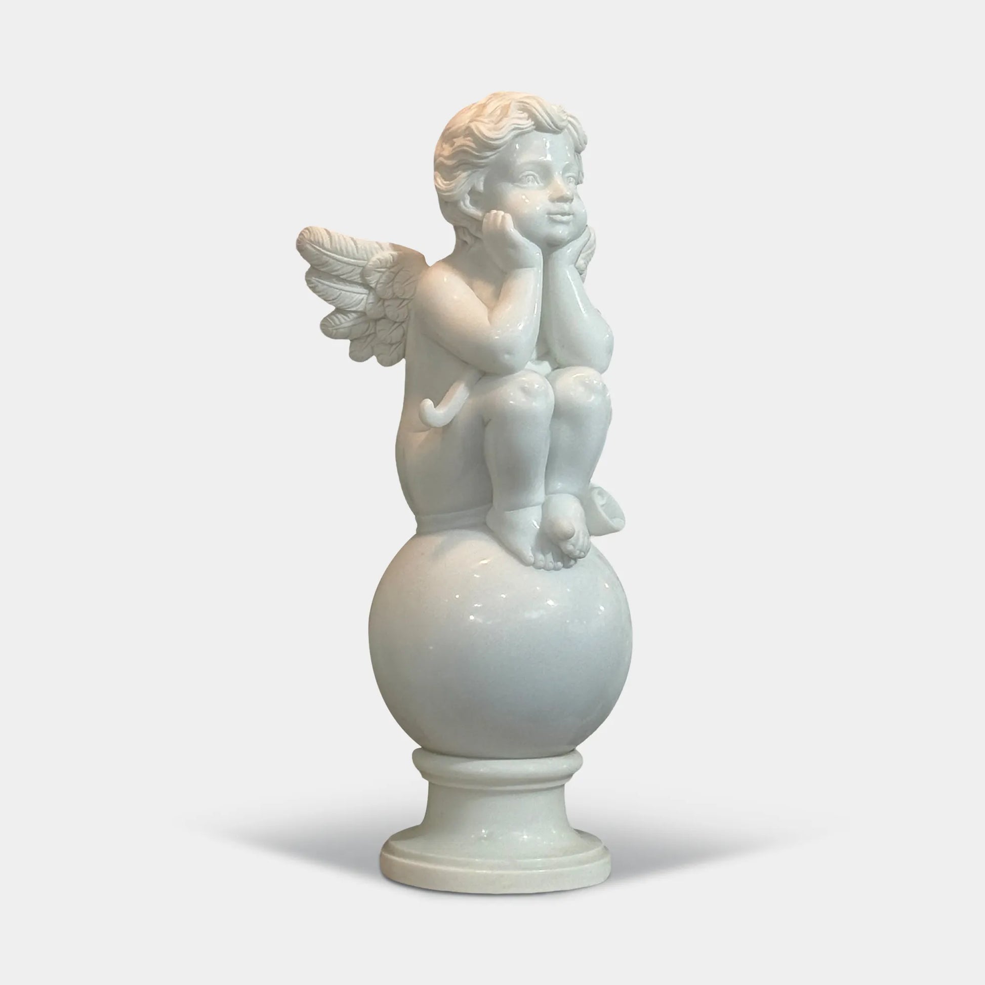 Angel cherub sitting with hands supporting face on a round stone pedestal carved in white marble in a pool center. 