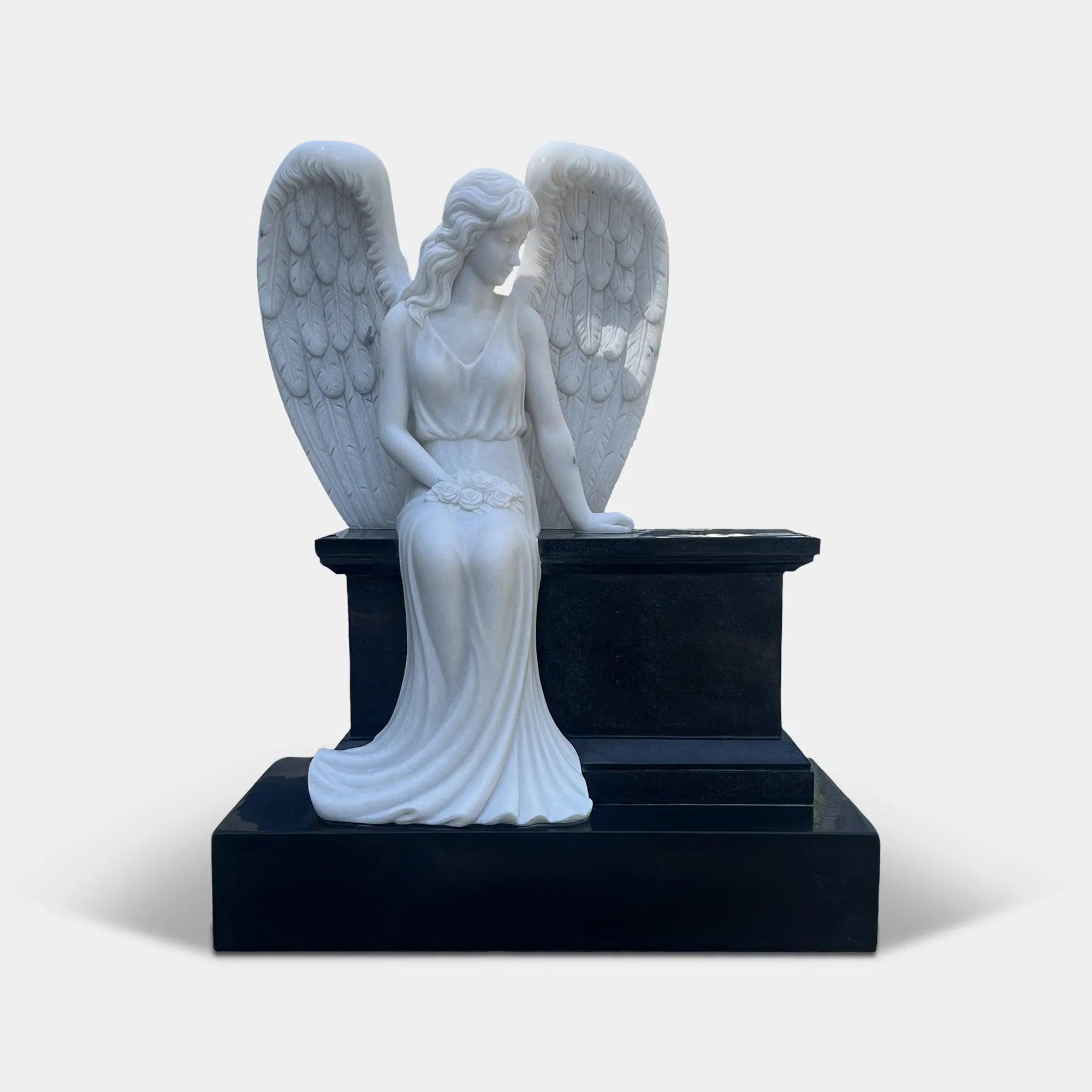 Marble angel statue seats on a black tombstone in a cemetery surrounded by foliage.