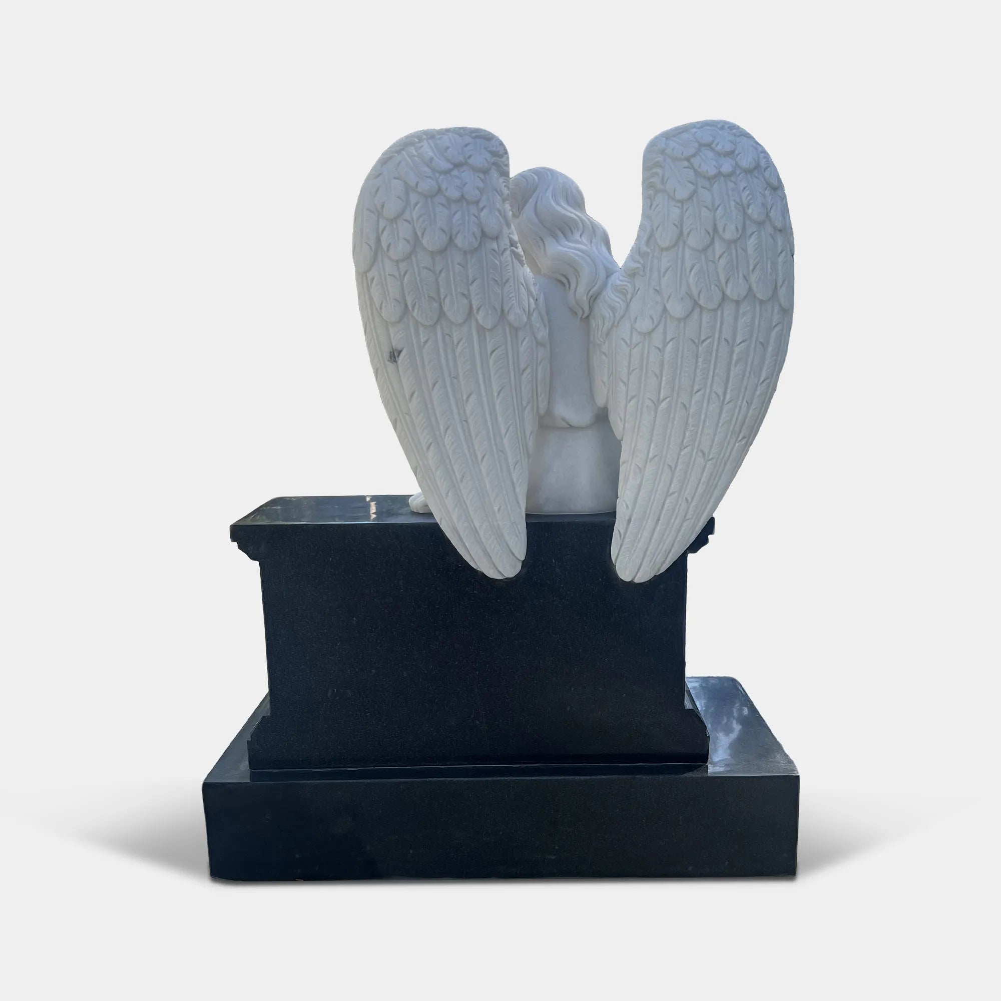 Marble angel statue seats on a black tombstone in a cemetery surrounded by foliage.