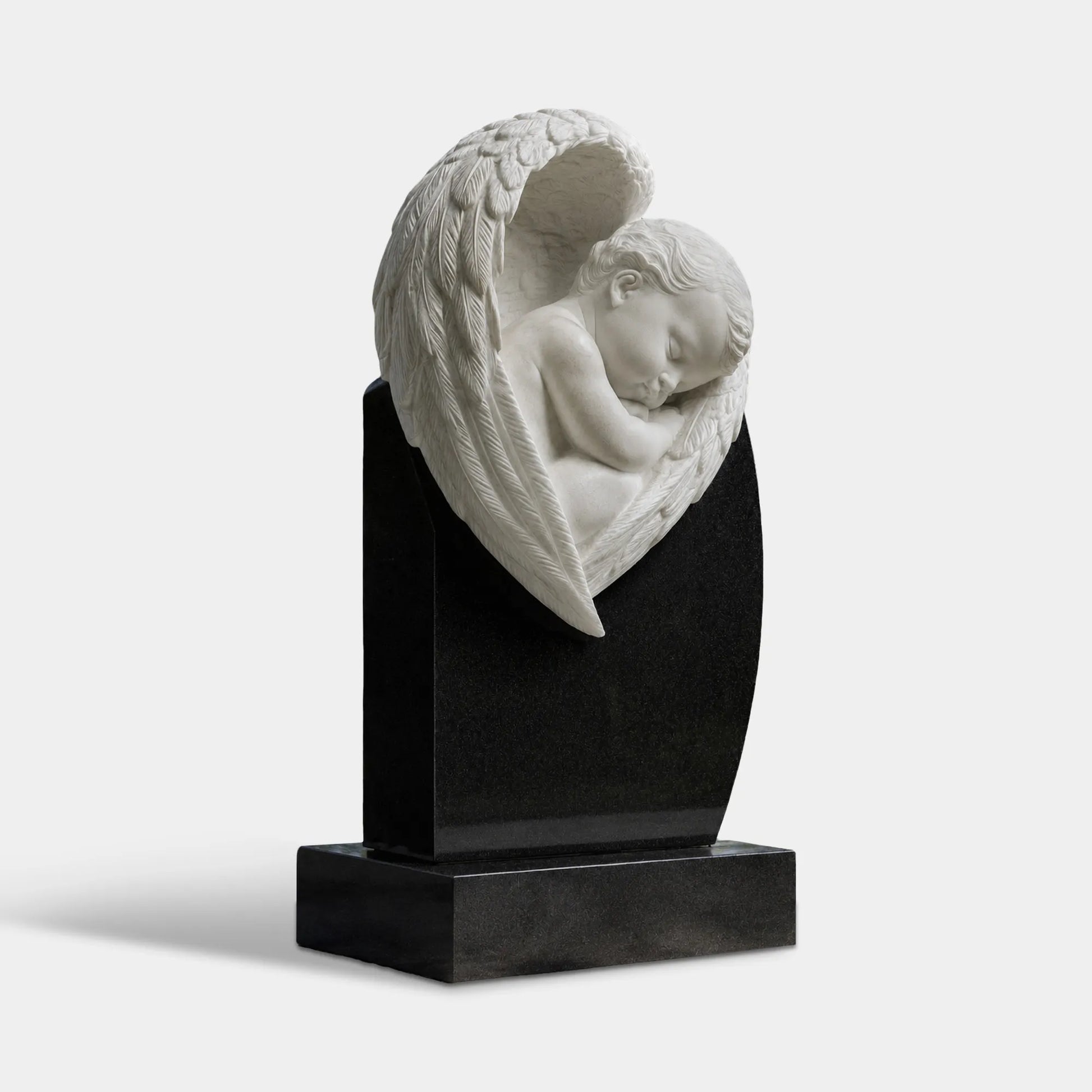 White marble sleeping child angel sculpture with one large wing mounted on a polished black marble headstone in a cemetery walkway.