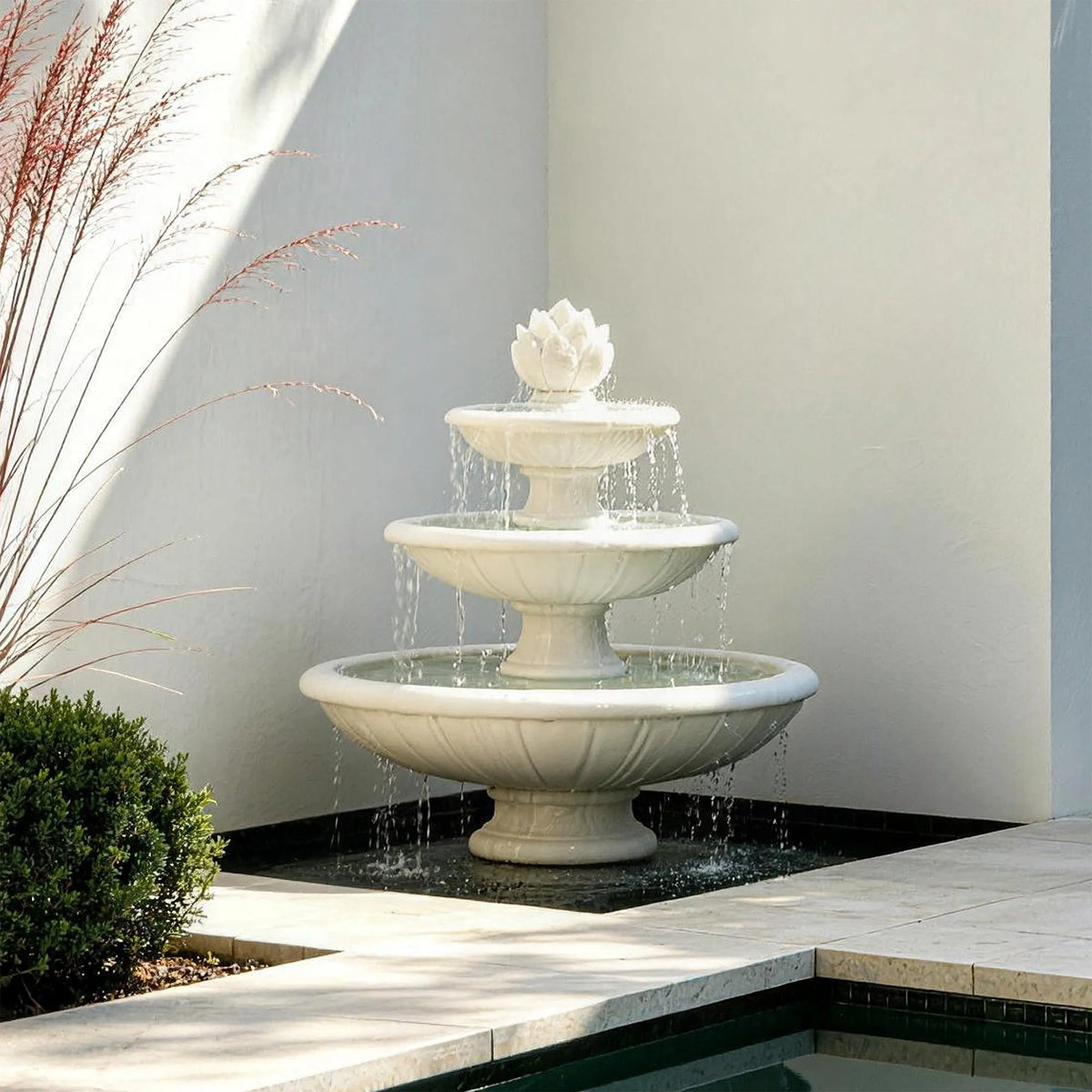 A three tier white marble fountain with lotus detail sits in a wall side outdoor patio space.