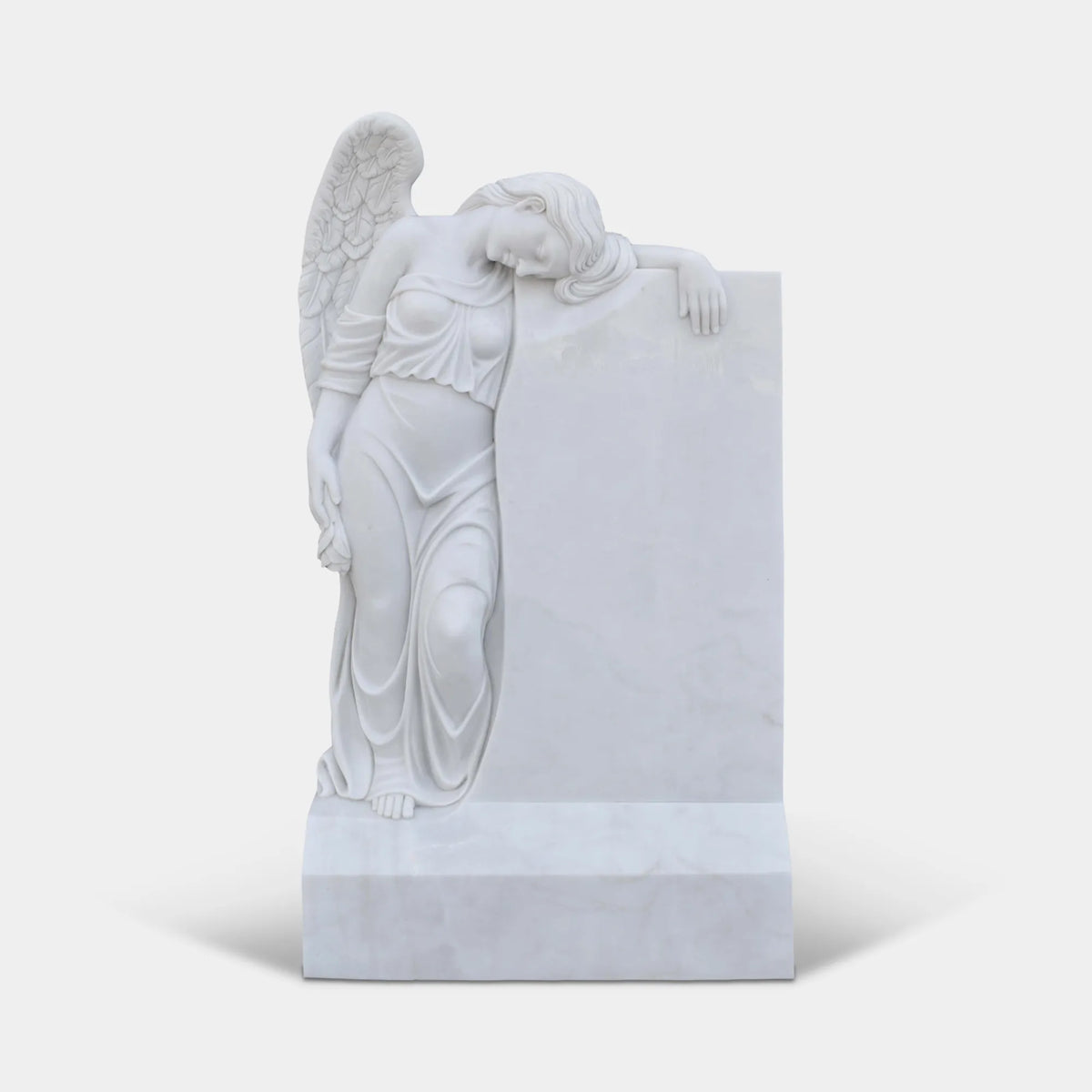 White marble grieving angel with detailed feathered wings, flowing gown, and bowed head leaning against a headstone in a cemetery garden.
