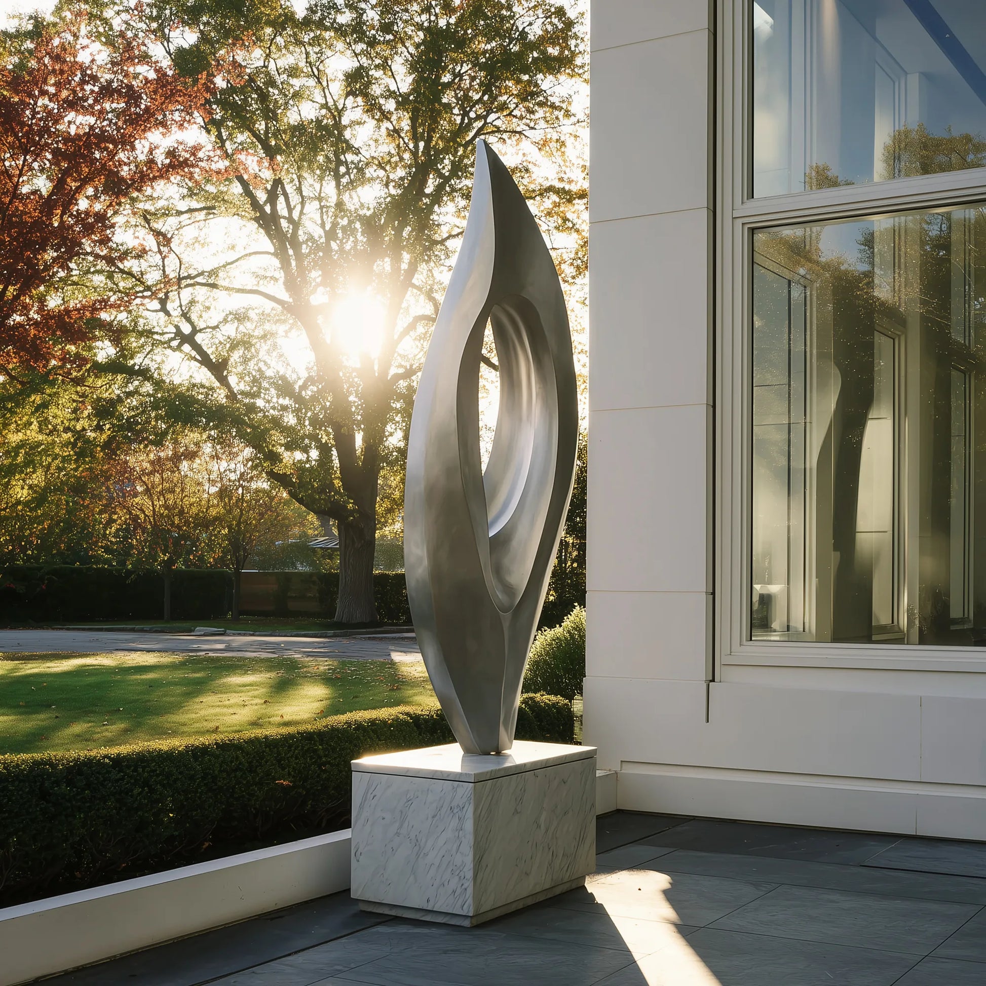 Abstract stainless steel sculpture with flowing hollow form on marble pedestal beside modern house exterior with autumn trees